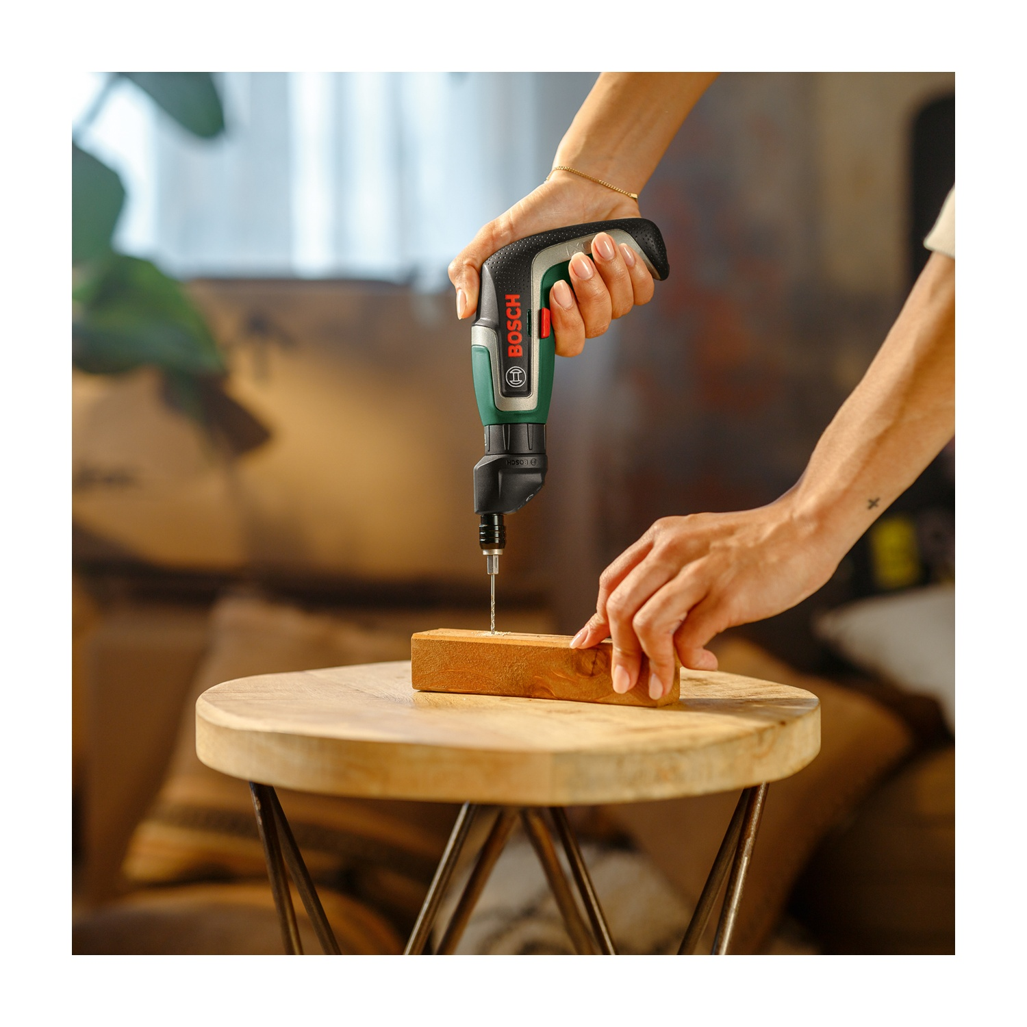 Mani che usano un trapano Bosch per forare il legno su un tavolo di legno. Il trapano è verde, nero e rosso. Lo sfondo è sfocato.