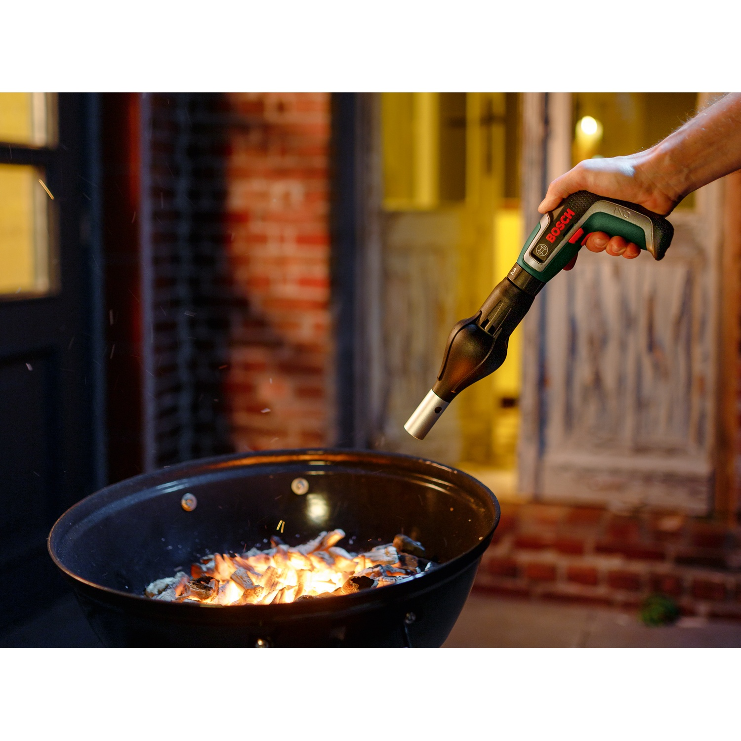 Una mano tiene un accendino Bosch per barbecue su una griglia a carbone con braci. Sfondo con muro di mattoni e porta di legno.