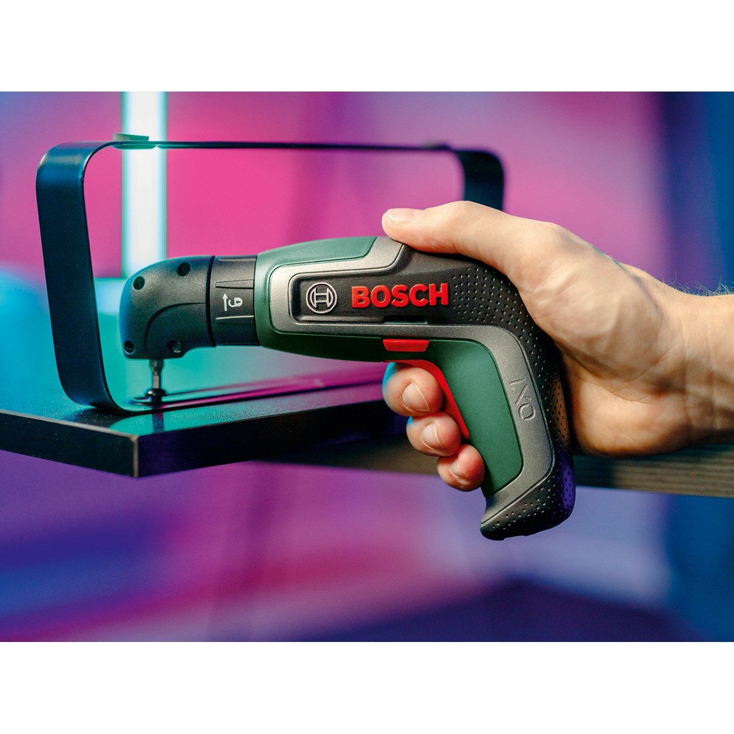Una mano tiene un cacciavite Bosch IXO verde e nero, vicino a un supporto in metallo nero, su uno sfondo sfocato e colorato.