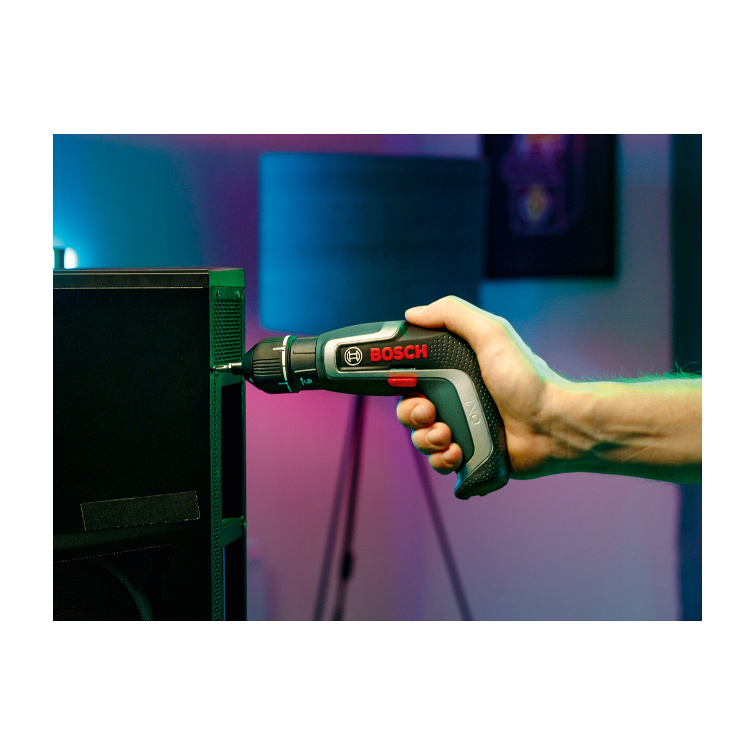La mano tiene un cacciavite a batteria Bosch IXO, che avvita un pannello nero. Lo sfondo ha un'illuminazione viola e blu sfocata.