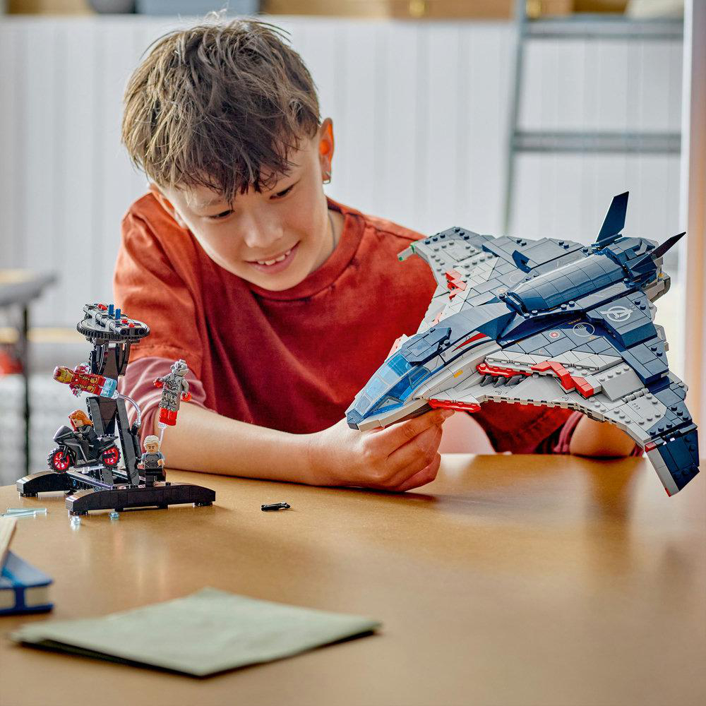 Ein Junge lächelt und hält ein großes Lego-Jet. Auf dem Tisch befinden sich ein Lego-Gebilde und Figuren.