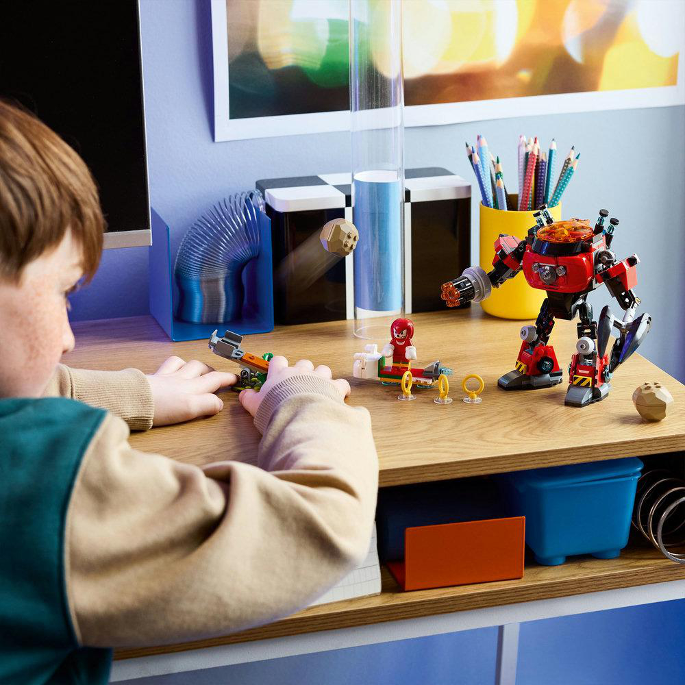Ein Kind spielt mit LEGO-Spielzeug auf einem Schreibtisch. Ein roter Roboter, ein grünes Fahrzeug und andere LEGO-Figuren sind sichtbar.