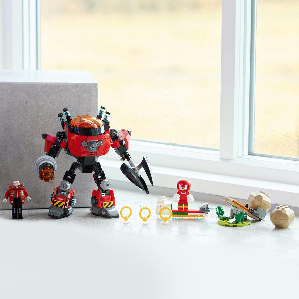 Ein Lego-Set mit einem roten Roboter, Figuren, Ringen und einer kleinen Szene. Der Roboter befindet sich im Vordergrund. Im Hintergrund ist ein Fenster.