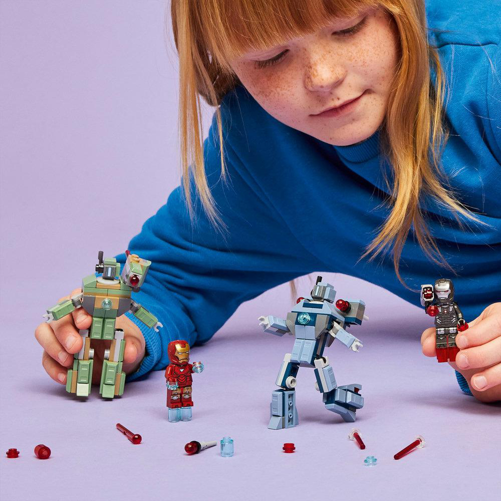 Ein Kind spielt mit Lego-Robotern und Iron-Man-Figuren auf einer lila Oberfläche. Zubehör ist verstreut. Das Kind trägt einen blauen Pullover.