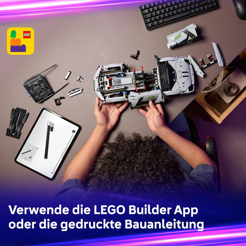 Hände halten ein LEGO-Automodell. Teile und Anweisungen sind verstreut. Tastatur und Tablet sind ebenfalls auf dem Tisch zu sehen.