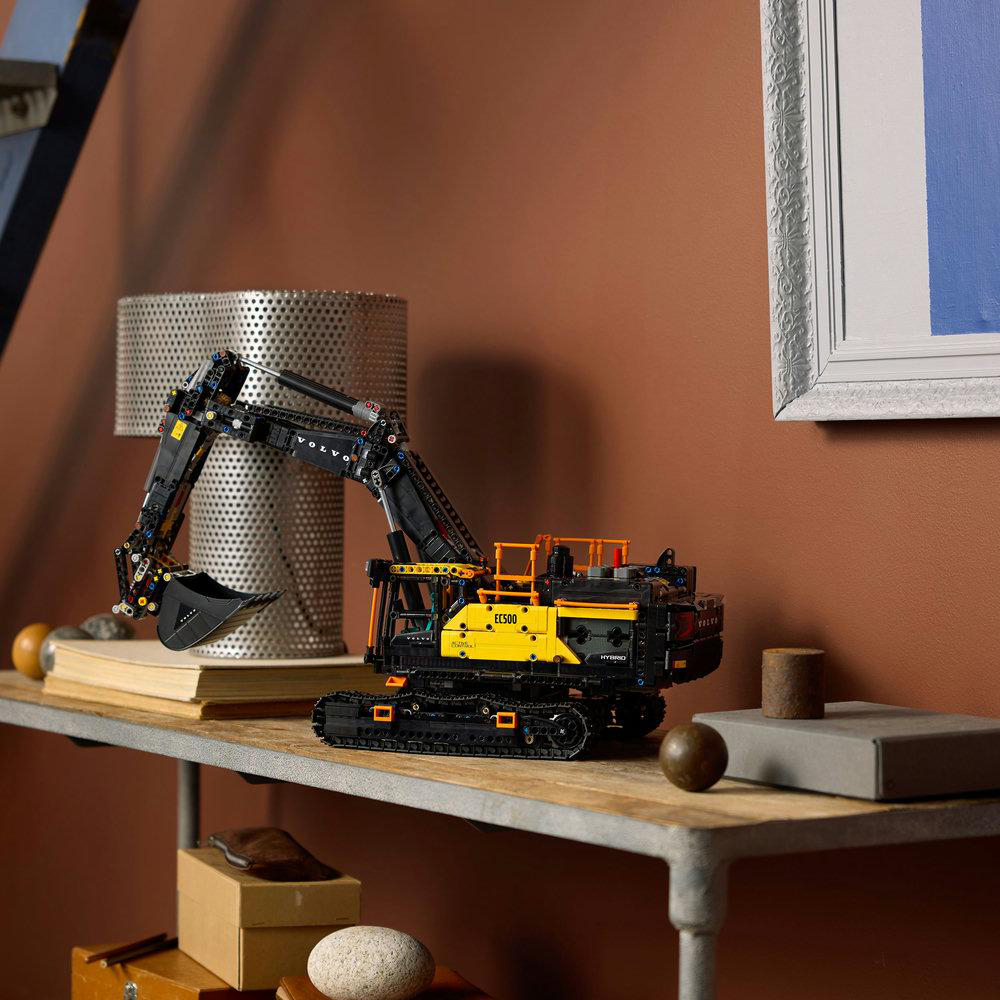 Ein Lego Volvo Bagger Modell auf einem Regal. Das Modell ist gelb und schwarz. Auf dem Regal befinden sich eine Lampe, Bücher und andere Gegenstände.