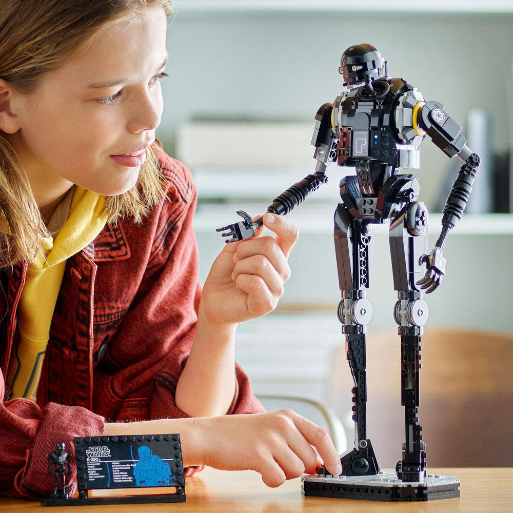 Ein Mädchen mit roter Jacke und gelbem Kapuzenpullover betrachtet eine schwarze Roboterfigur. Ein Star Wars Lego-Set steht auf dem Tisch.