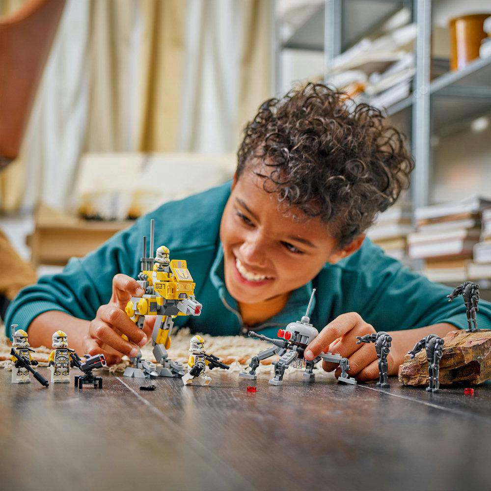 Ein lächelnder Junge spielt mit LEGO Star Wars Figuren. Ein gelber Walker, Droiden und Minifiguren sind auf dem Holzboden.