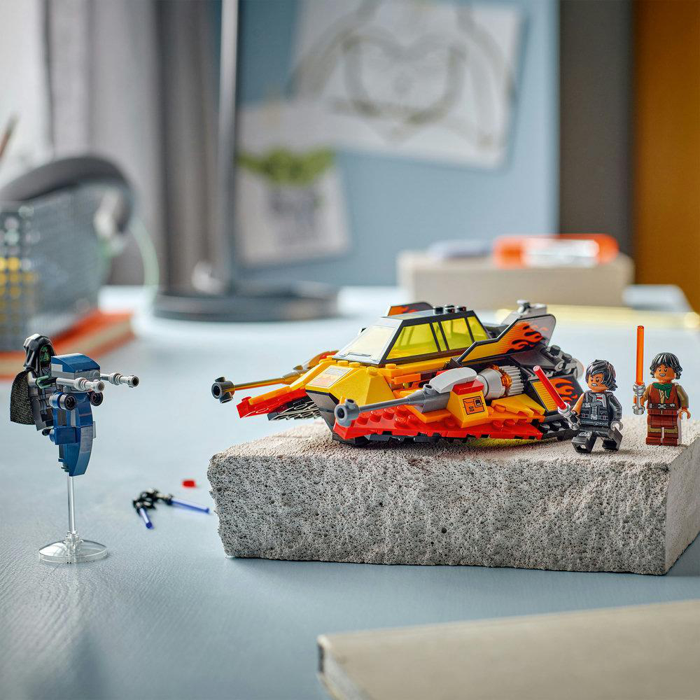 Lego-Set. Ein gelb-rotes Raumschiffmodell steht auf einem Block. Lego-Figuren stehen daneben. Eine blaue Lego-Figur links.