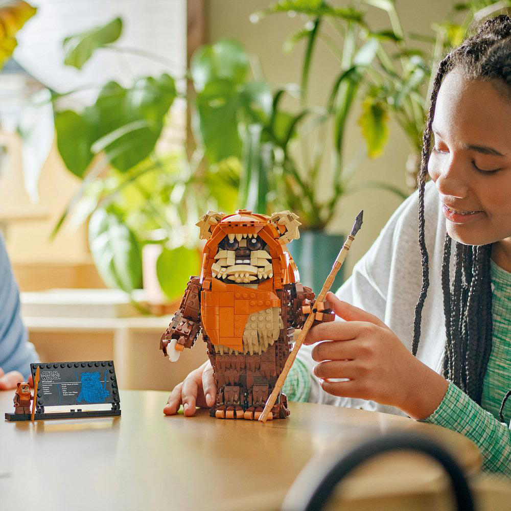Ein Mädchen mit einer Lego-Ewok-Figur, die einen Speer hält. Die Figur ist braun und orange. Im Hintergrund ist eine Pflanze zu sehen.