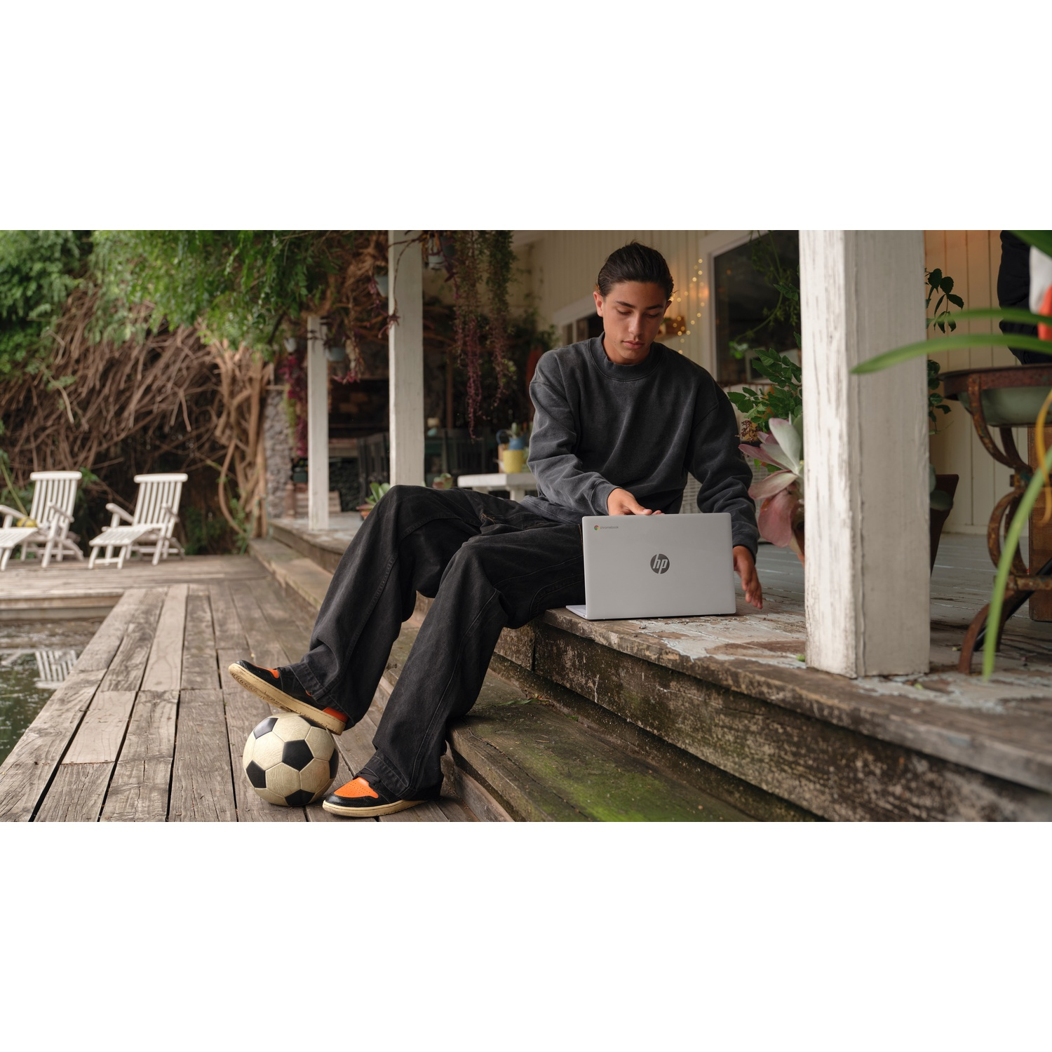 A person sits with a laptop and a soccer ball on wooden steps. The background shows a porch and greenery.