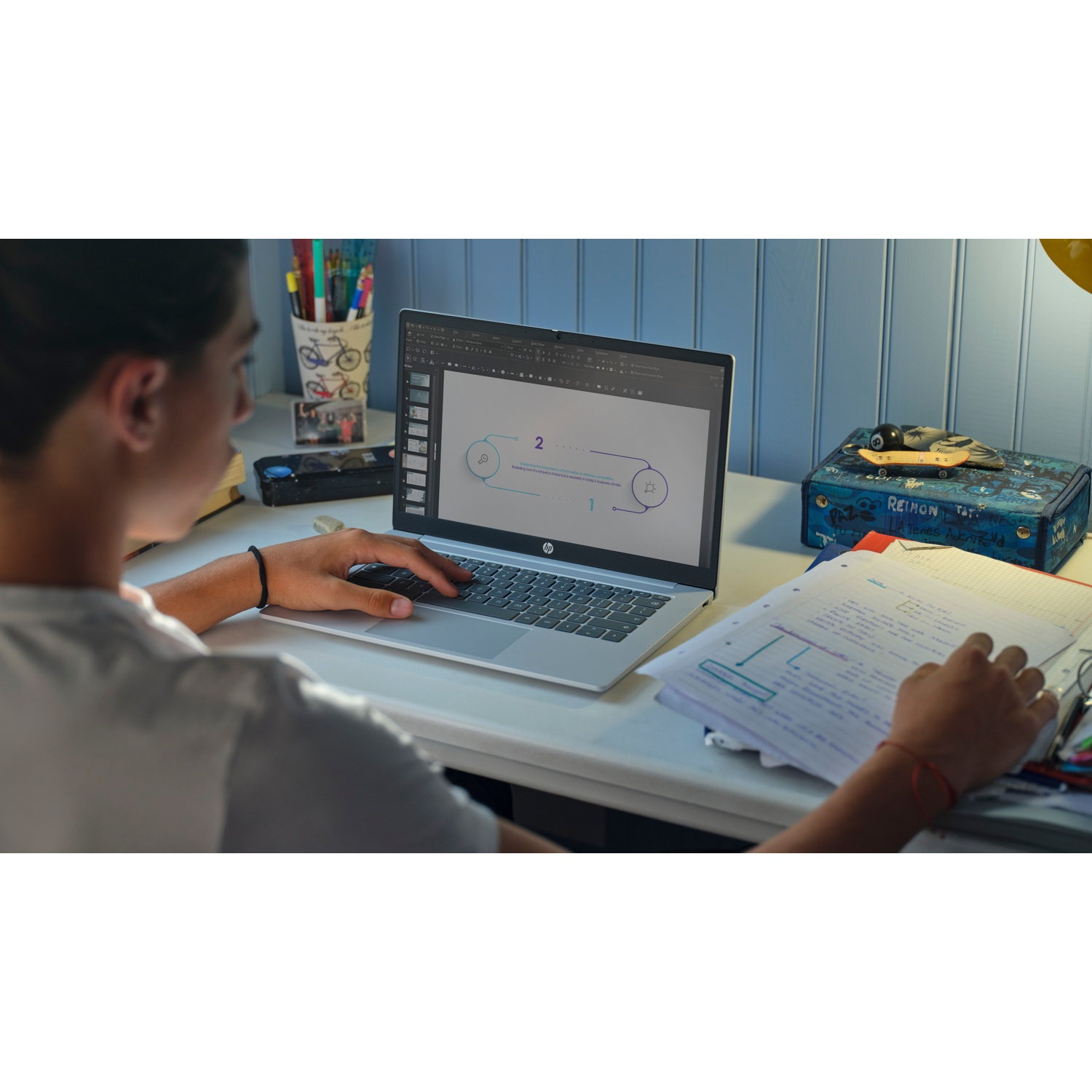 A person using a laptop, studying. There is a notebook with notes, a pencil holder, and a decorated box on the desk.