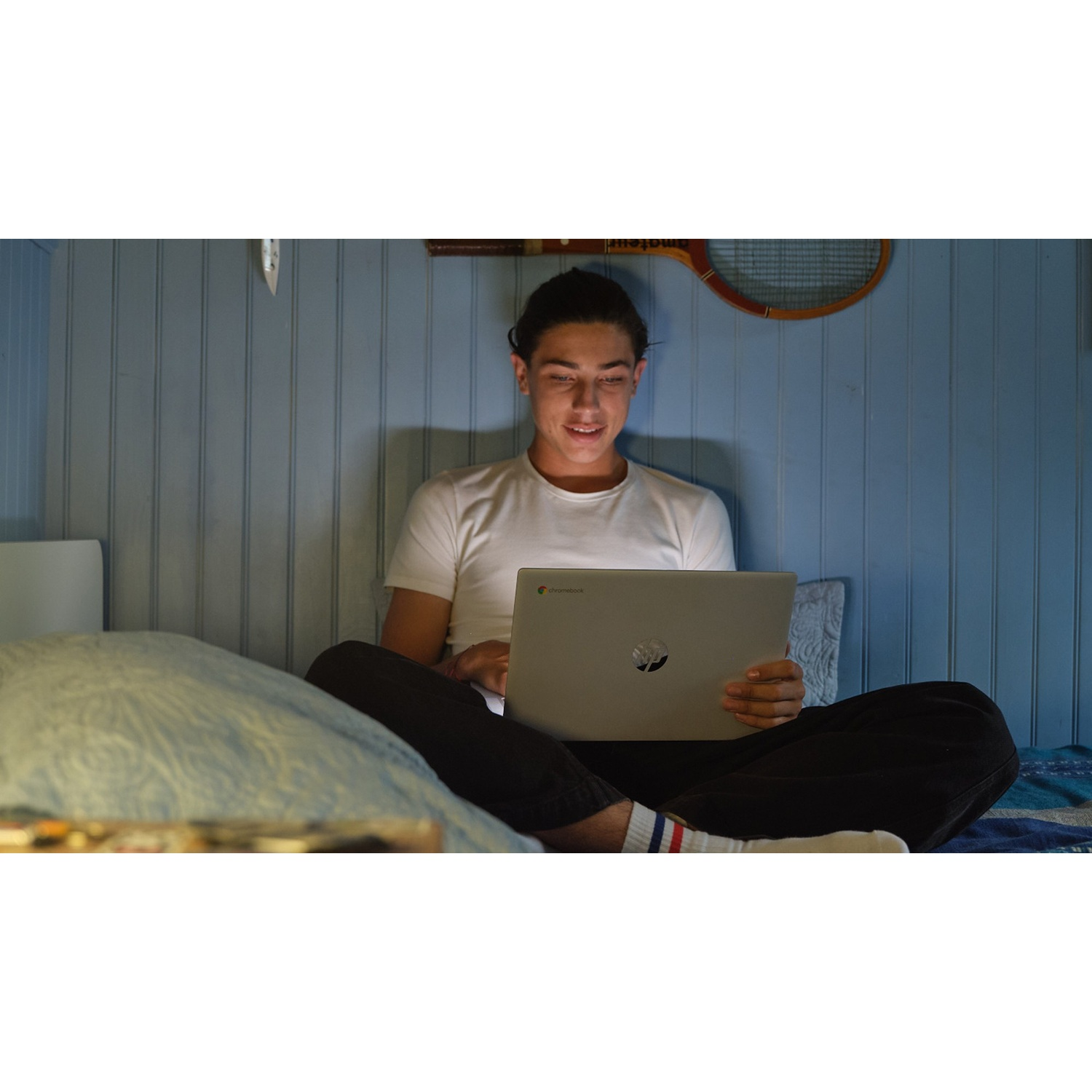 A young man sits on a bed, using a laptop. He wears a white tee and black pants. The room has blue walls and a tennis racket.