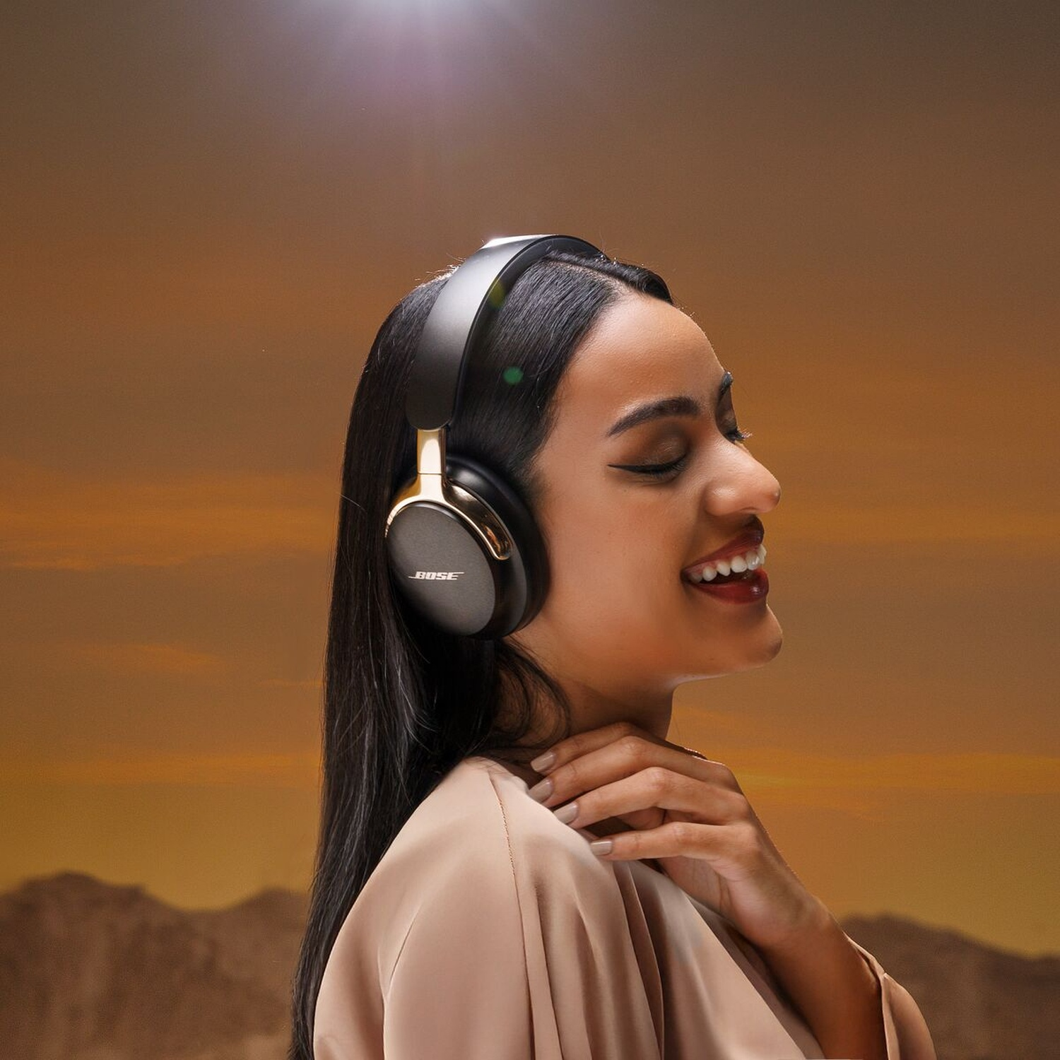 Une femme portant un casque Bose noir, souriante, devant un ciel orange. Elle se touche le cou et a de longs cheveux noirs.