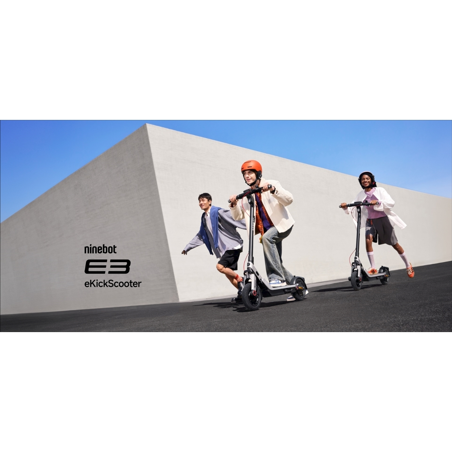 Tre persone guidano e-scooter su una superficie pavimentata. Un edificio grigio e un cielo blu sullo sfondo. Il logo 'ninebot E3 eKickScooter' è sul muro.