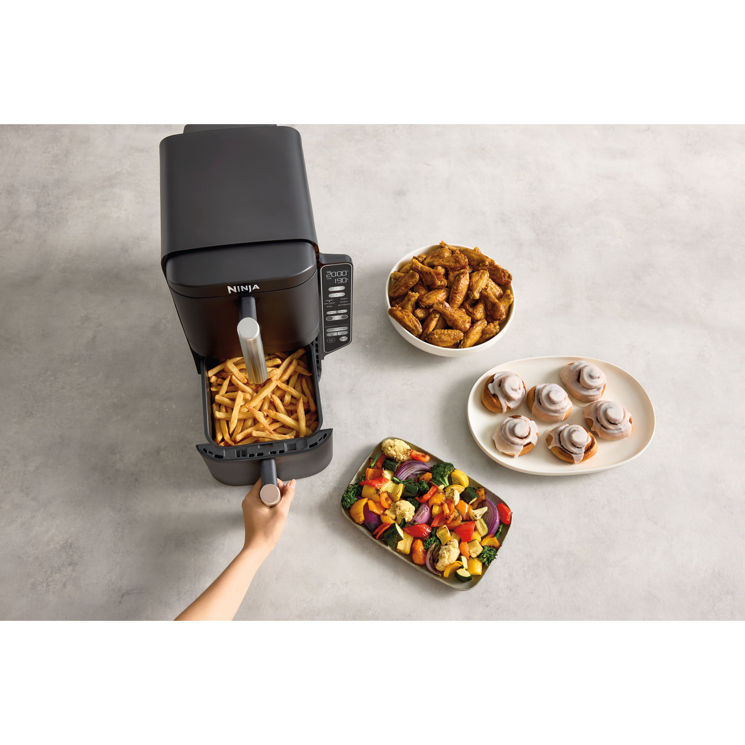 Black air fryer with fries, wings, and cinnamon rolls. Vegetables are also present. Hand opening the air fryer.