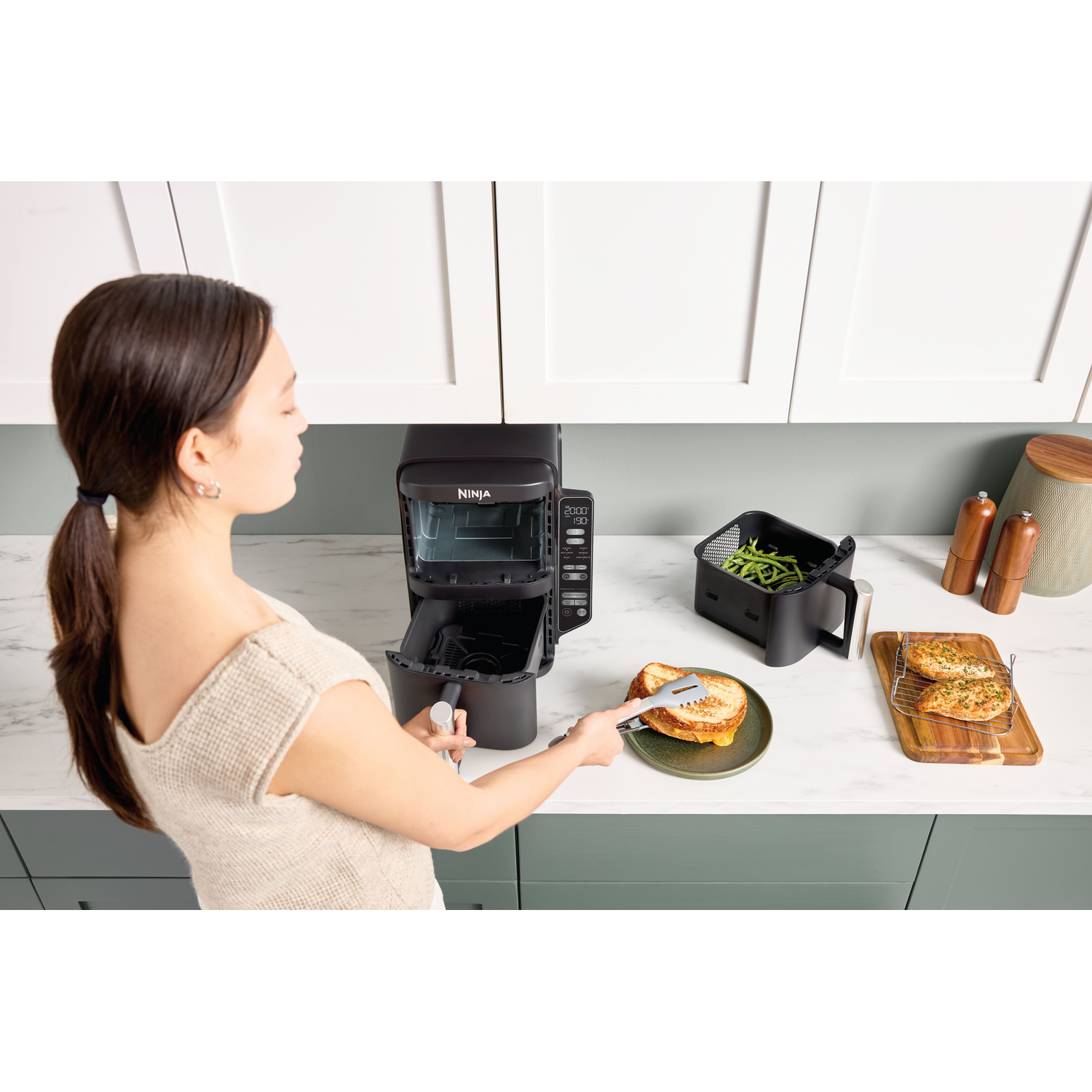 A woman uses tongs to take a sandwich from a plate. There is an air fryer, a tray with chicken, and a basket with beans.