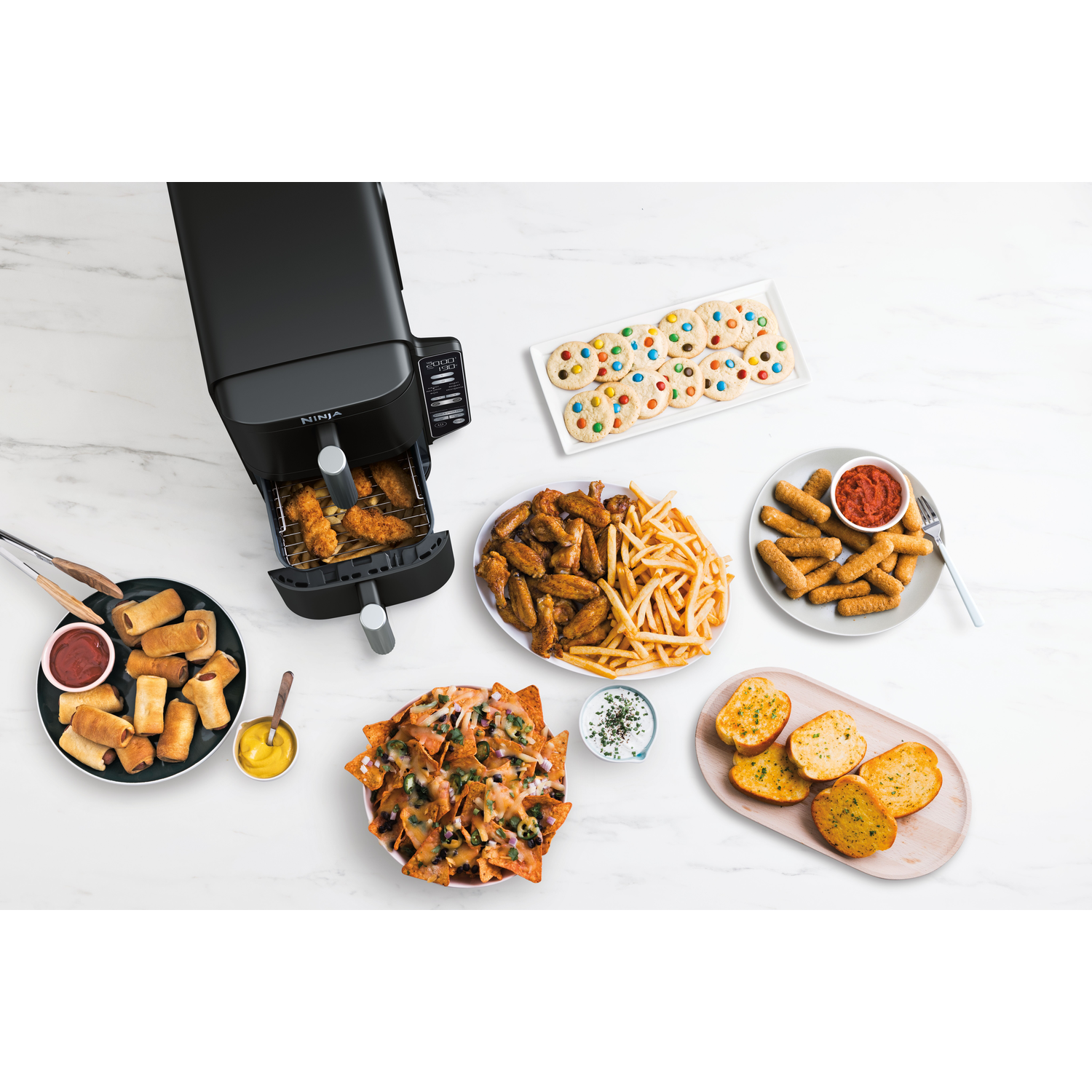 An air fryer with chicken wings, fries, nachos, cookies, mozzarella sticks, garlic bread, and pigs in a blanket on a white marble surface.