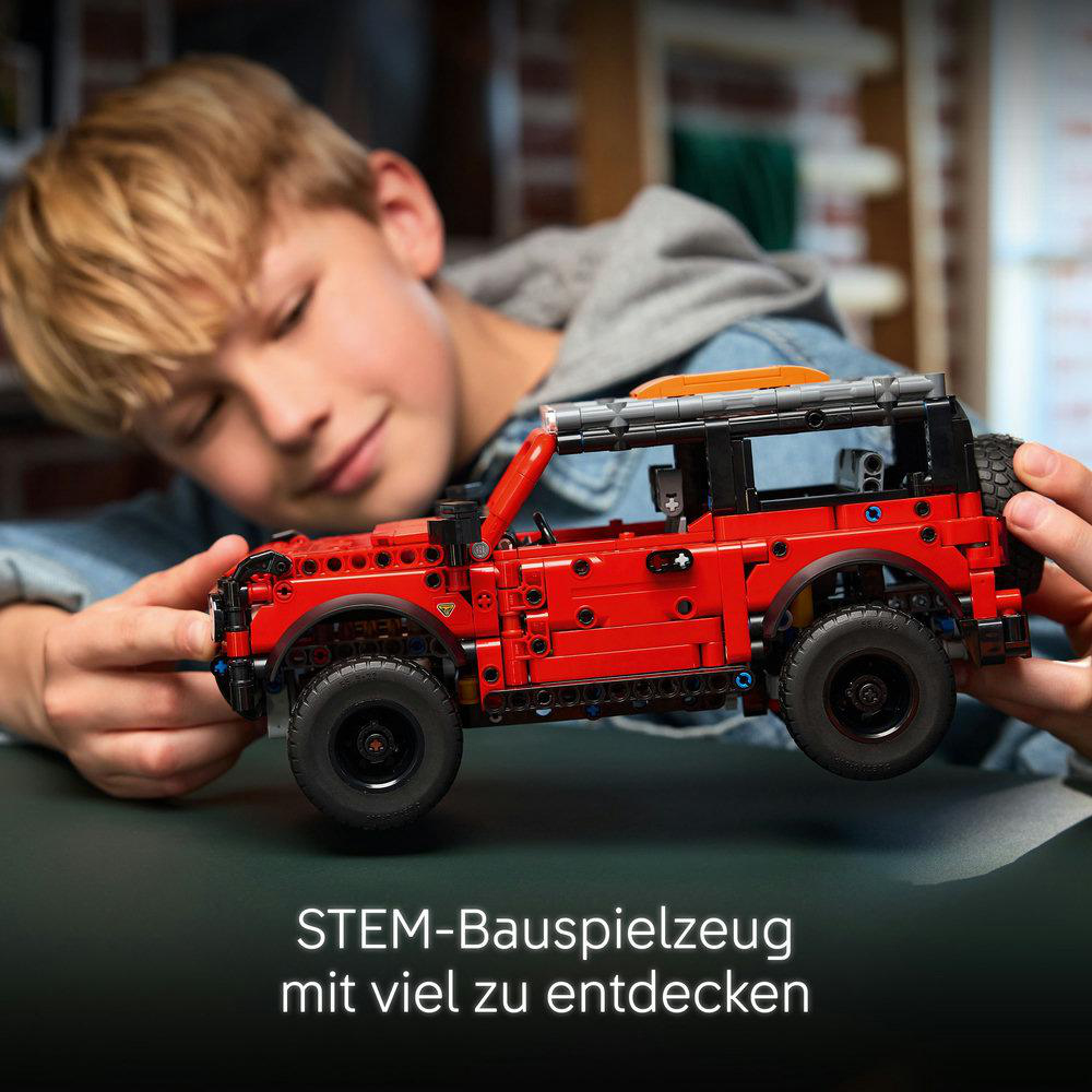 Ein Junge hält ein rotes Spielzeugauto. Das Auto ist detailliert und hat schwarze Räder. Der Junge ist blond und trägt eine Jeansjacke.