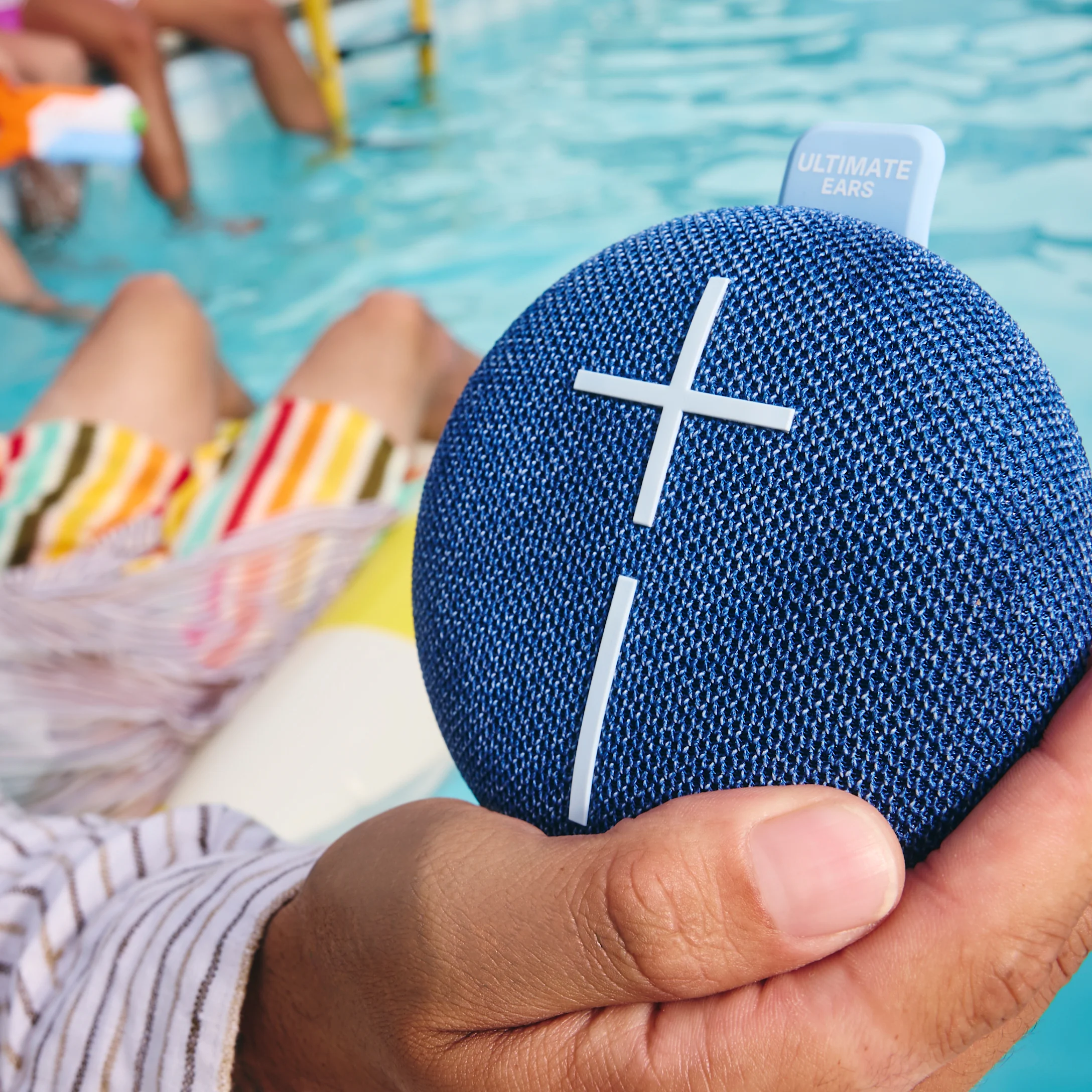 Eine Hand hält einen blauen UE-Lautsprecher. Die Szene spielt an einem Pool mit Beinen im Wasser.