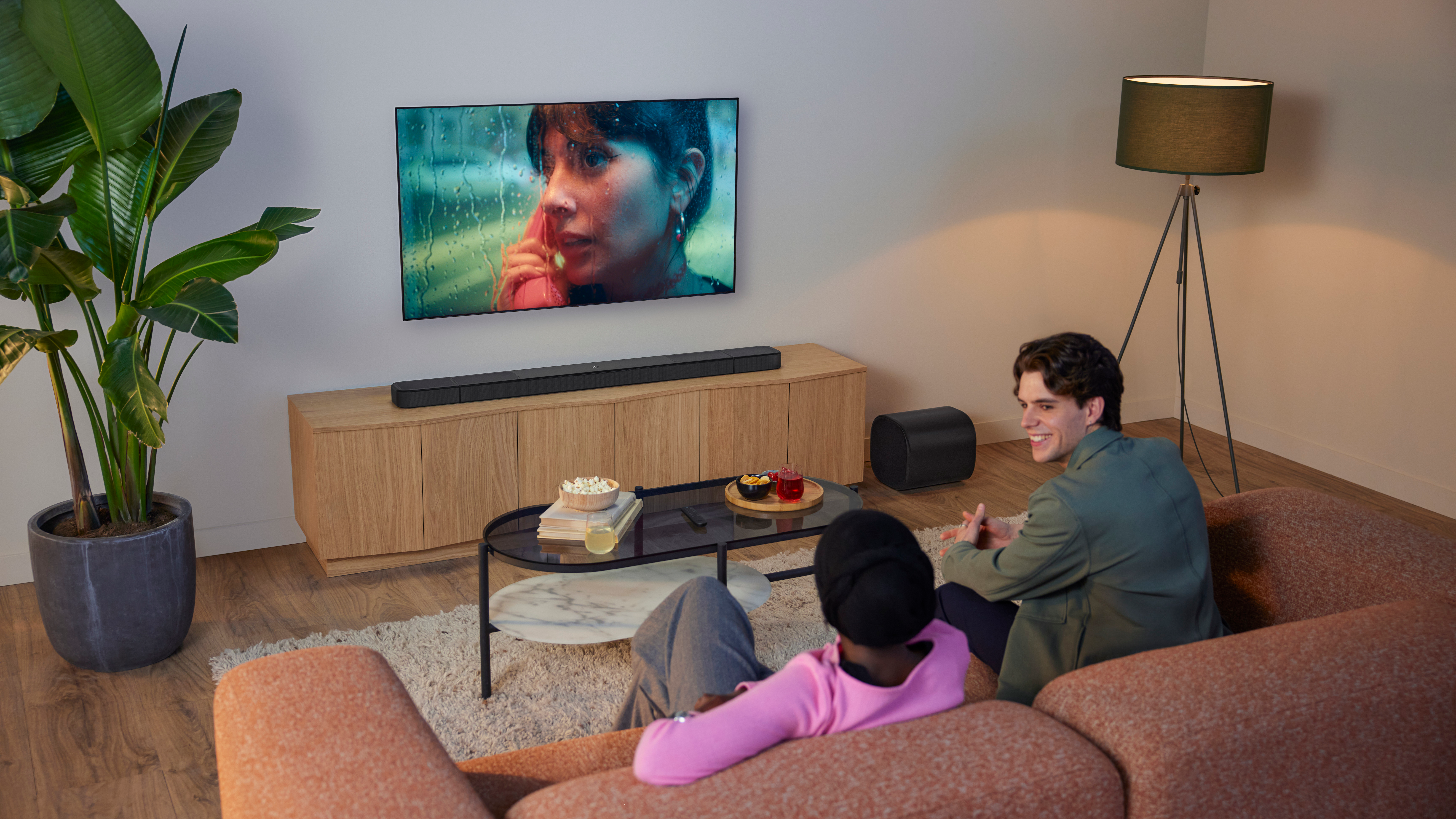 Wohnzimmerszene mit zwei Personen auf dem Sofa, die fernsehen. Es gibt eine Pflanze, eine Lampe und einen Couchtisch mit Snacks.
