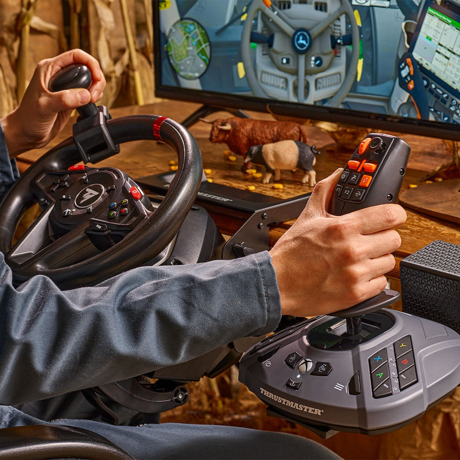 Eine Person benutzt ein Thrustmaster-Lenkrad und einen Joystick. Auf einem Bildschirm ist ein Spiel mit Kühen zu sehen. Die Person trägt ein graues Hemd.