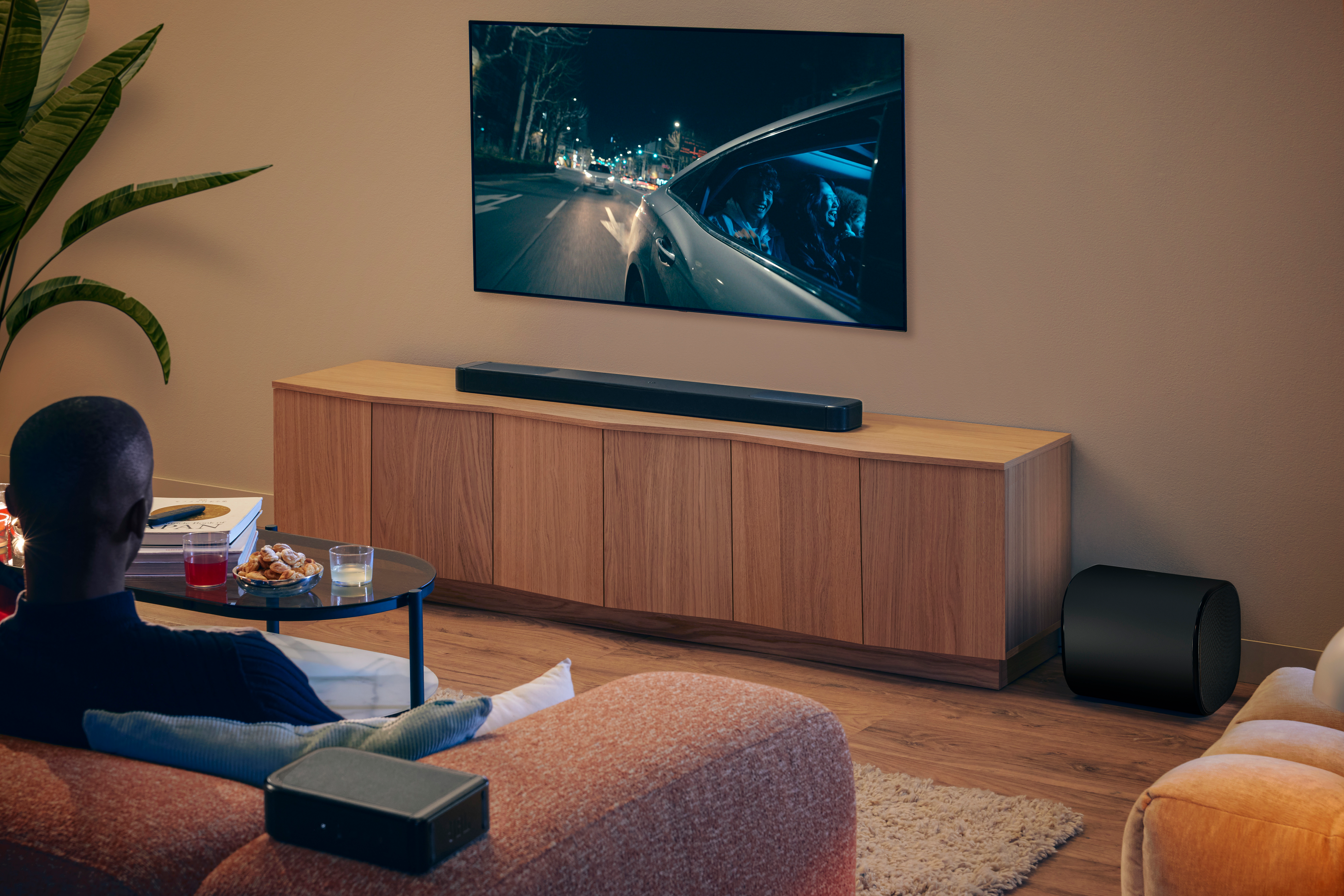 Eine Person schaut fern, auf dem ein Auto bei Nacht zu sehen ist. Soundbar, Subwoofer und Schrank sind sichtbar. Snacks und Getränke auf dem Tisch.