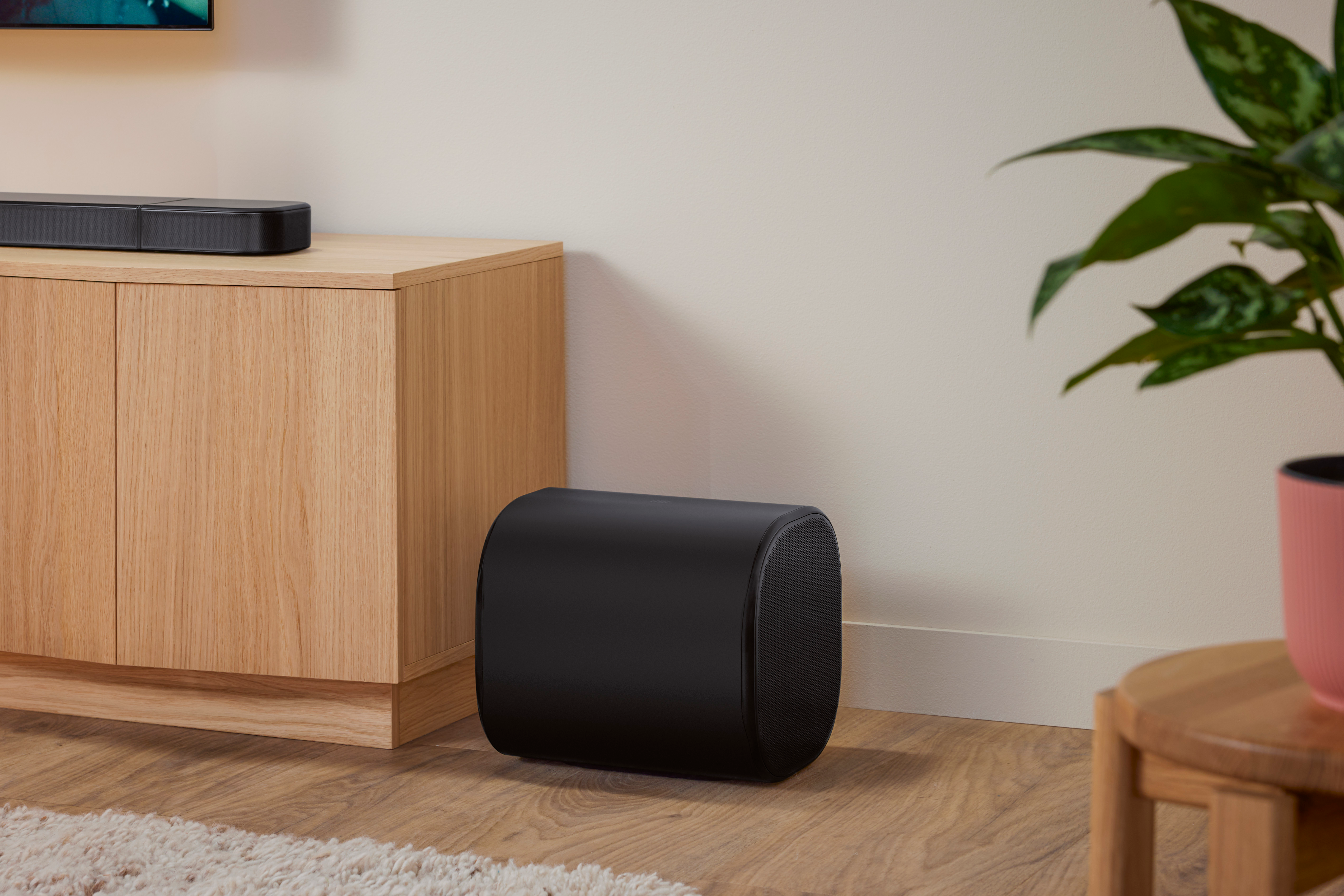Schwarzer Subwoofer auf Holzboden. Ein Holzschrank und eine Soundbar befinden sich links. Rechts ist eine Pflanze.