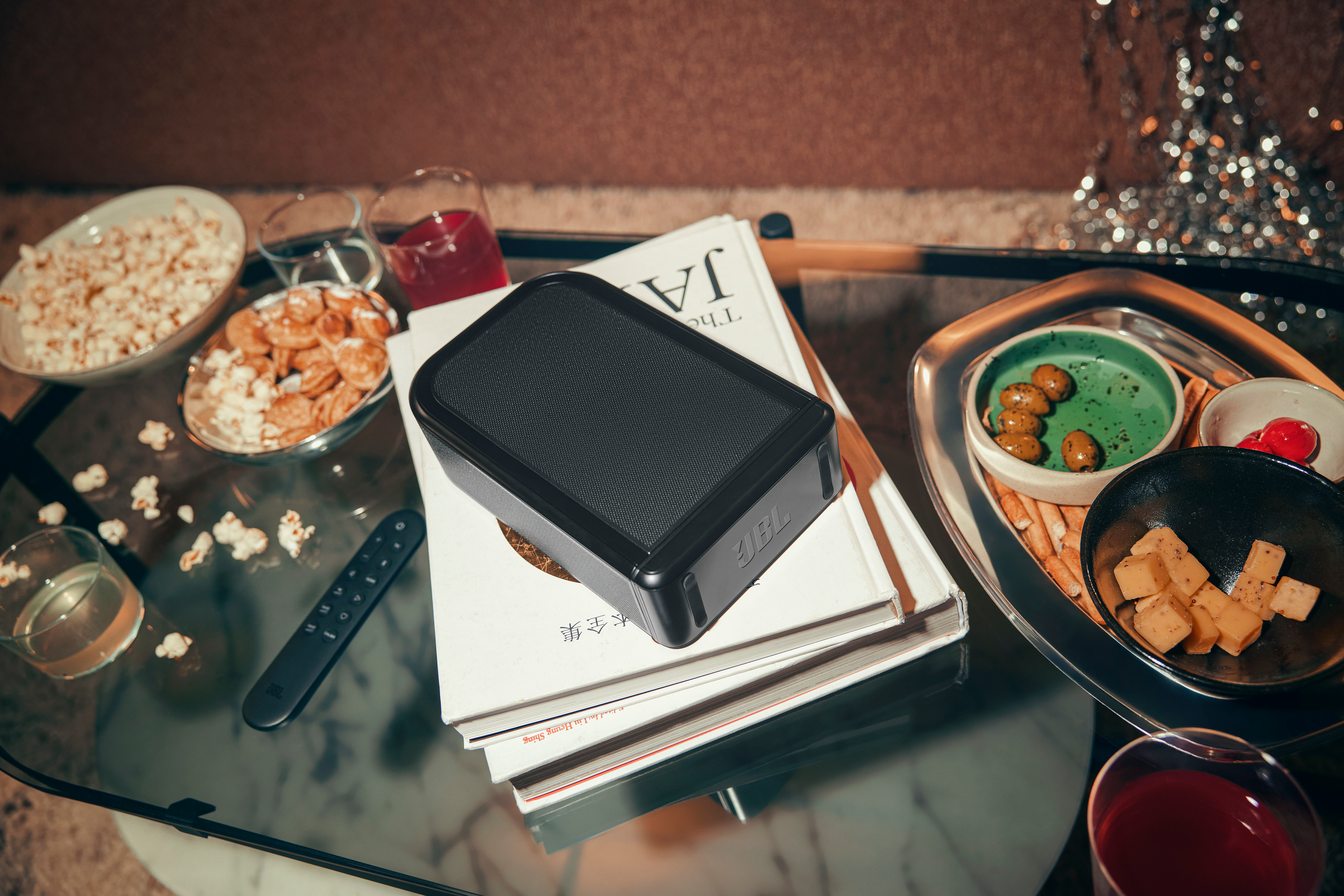 Ein schwarzer JBL-Lautsprecher auf Büchern. Snacks wie Popcorn, Oliven und Würfel liegen auf dem Glastisch. Getränke und eine Fernbedienung sind vorhanden.