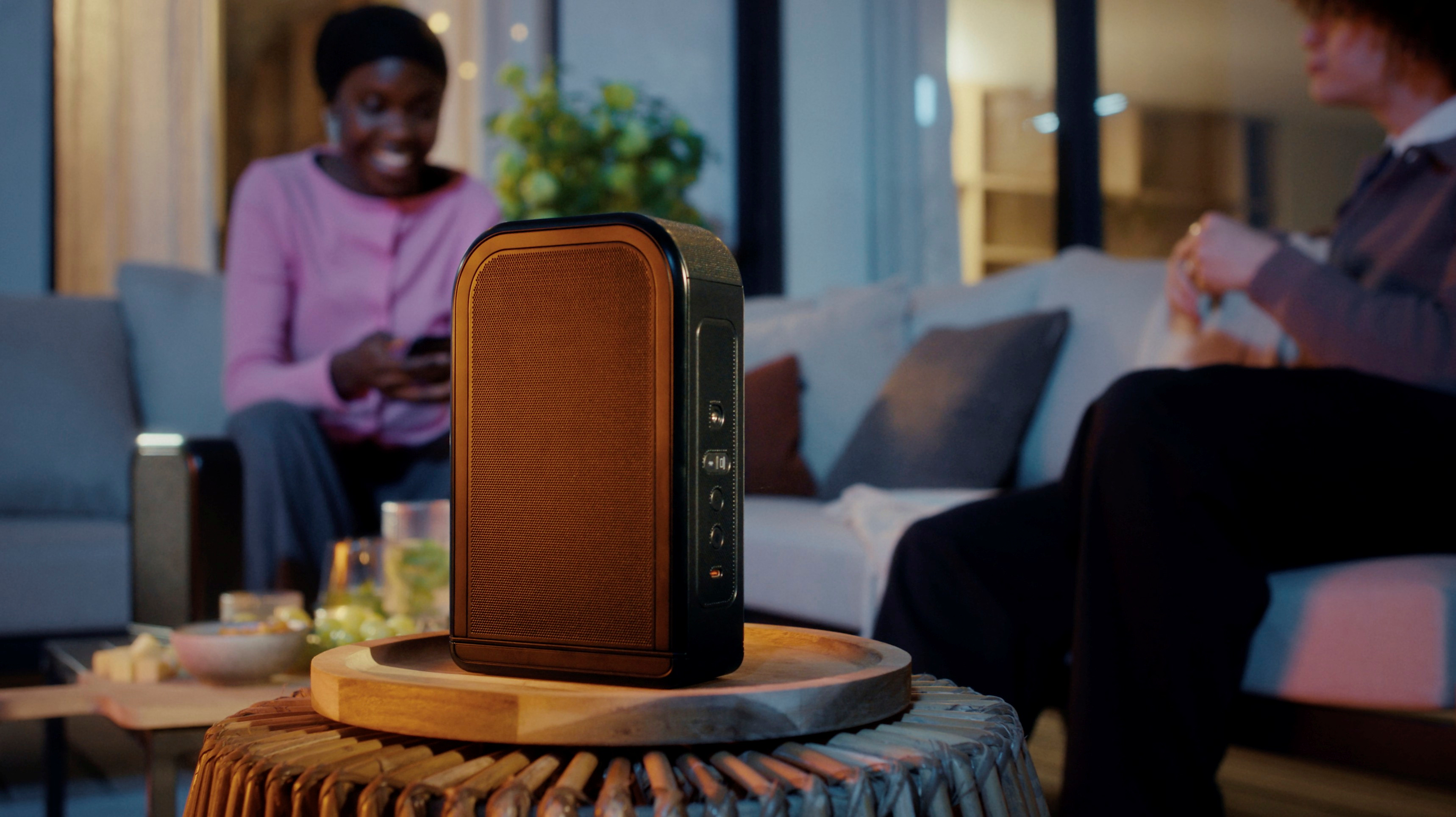 Ein Lautsprecher steht auf einem Tisch vor Personen auf einem Sofa. Eine Person hält ein Telefon. Snacks und Getränke sind in der Nähe.