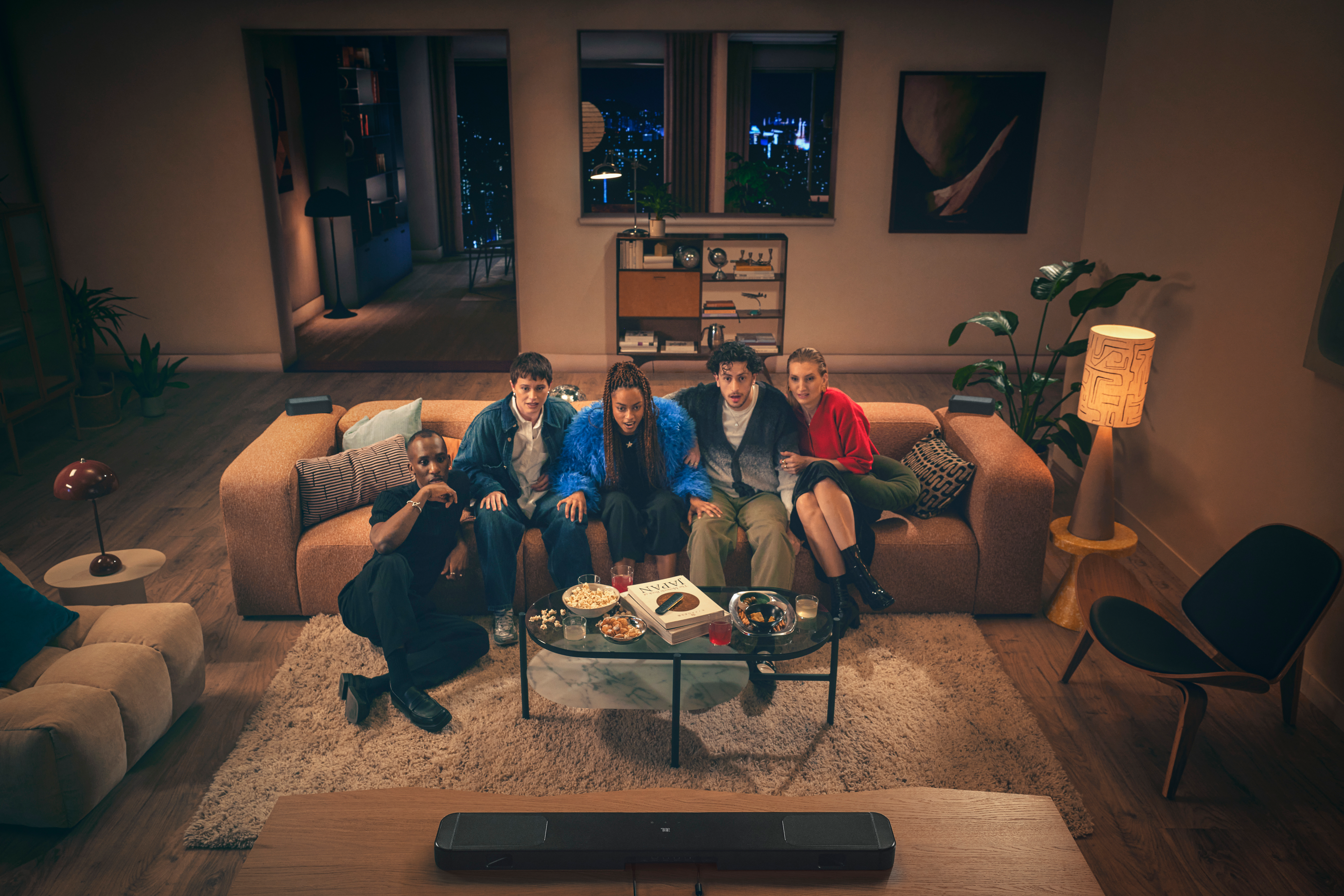 Fünf verschiedene Personen sitzen in einem Wohnzimmer auf einem Sofa und schauen etwas. Ein Tisch mit Snacks steht vor ihnen. Stadtblick.