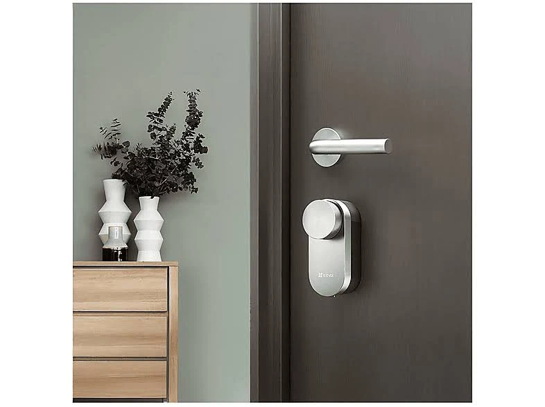 Una puerta moderna con manija plateada y cerradura inteligente. A la izquierda, una cómoda de madera con jarrones y plantas.
