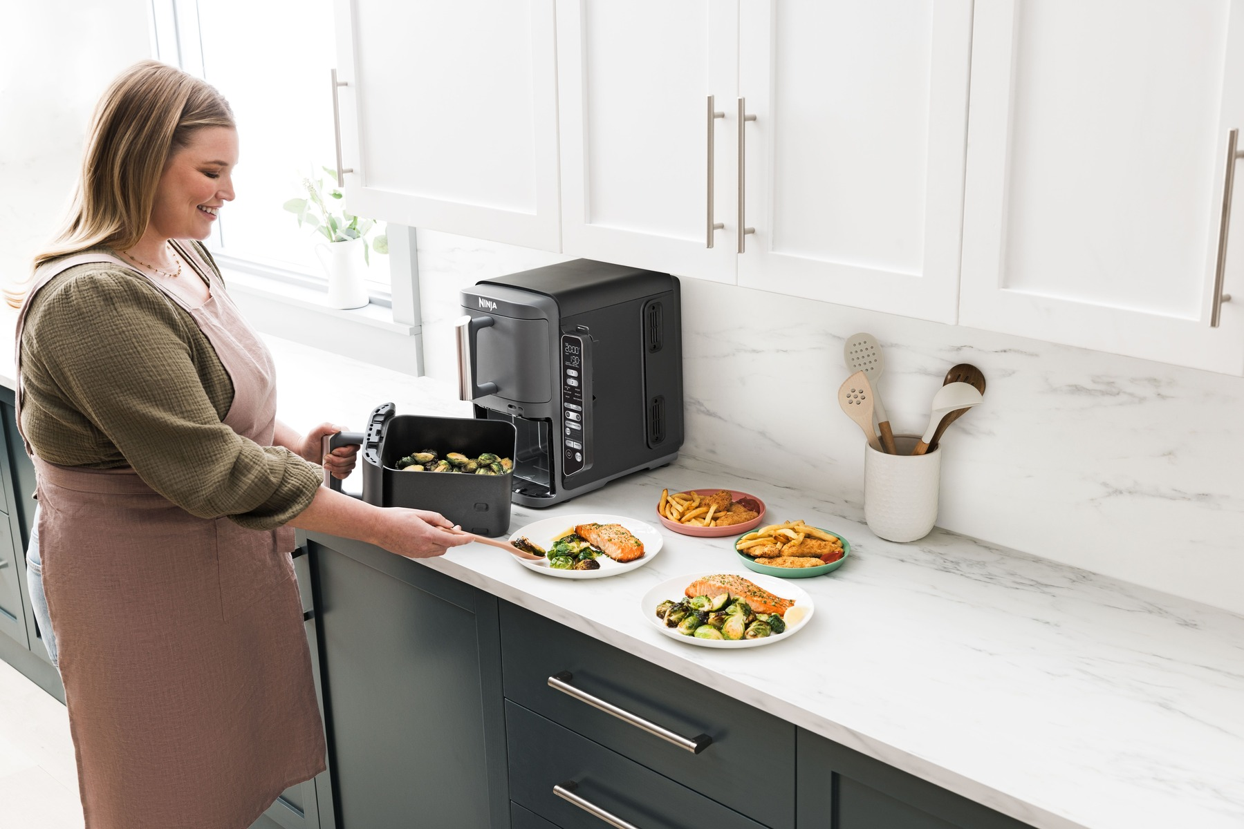 Una mujer con delantal cocina en una cocina. Varios platos de comida están en la encimera, con una freidora de aire en el fondo.
