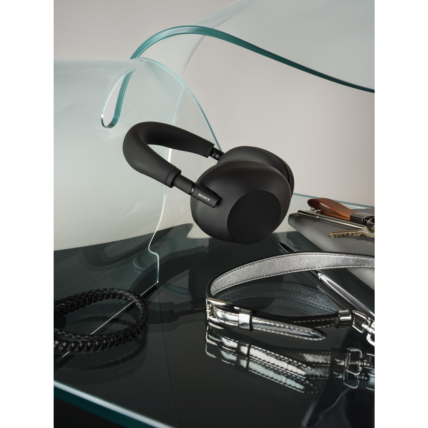 Zwarte Sony koptelefoon, armband, riem, sleutels en notitieboek op een reflecterend oppervlak, met een gebogen glazen structuur op de achtergrond.