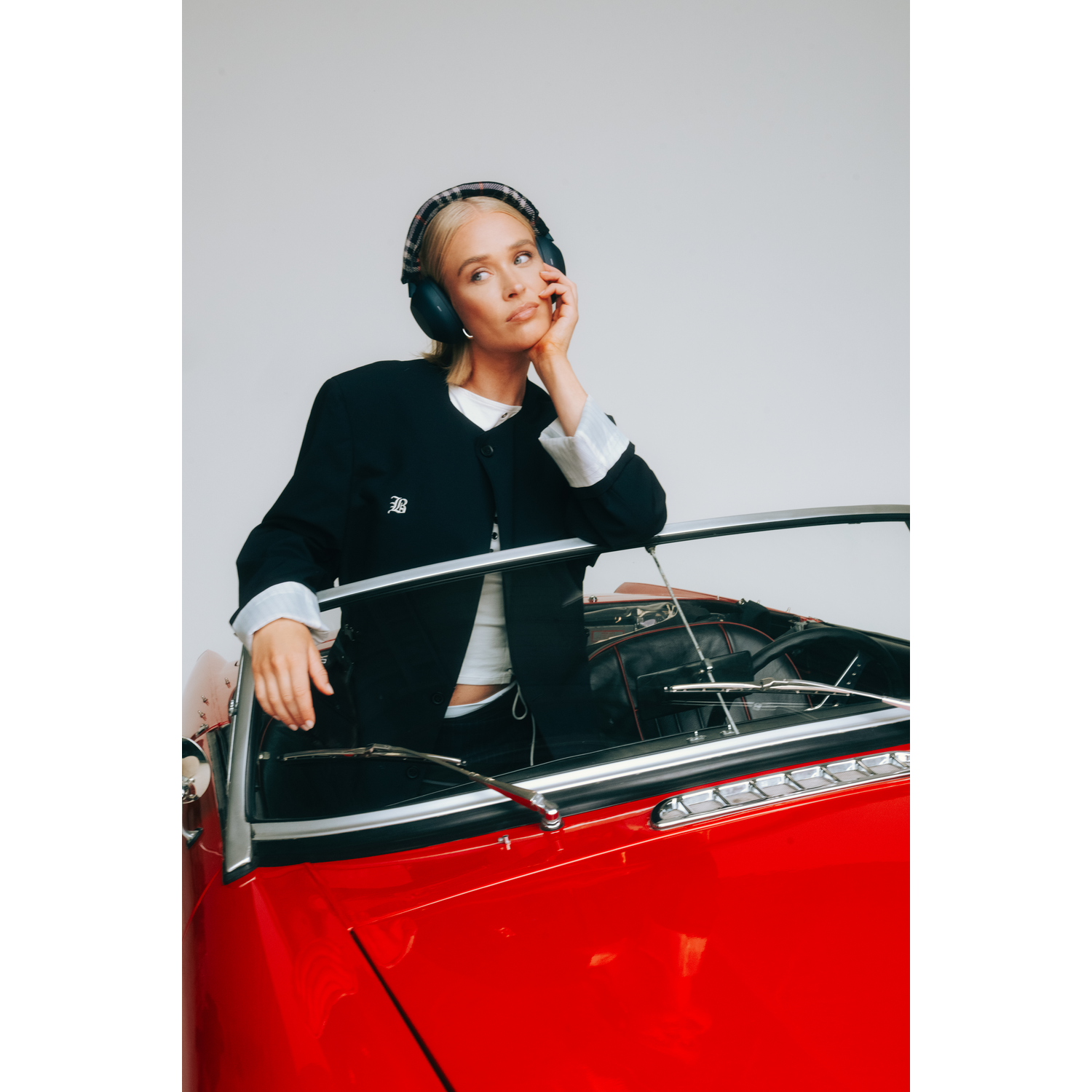 Een vrouw in een zwarte jas en koptelefoon leunt op een rode auto. Ze rust haar hand op haar wang en kijkt nadenkend.