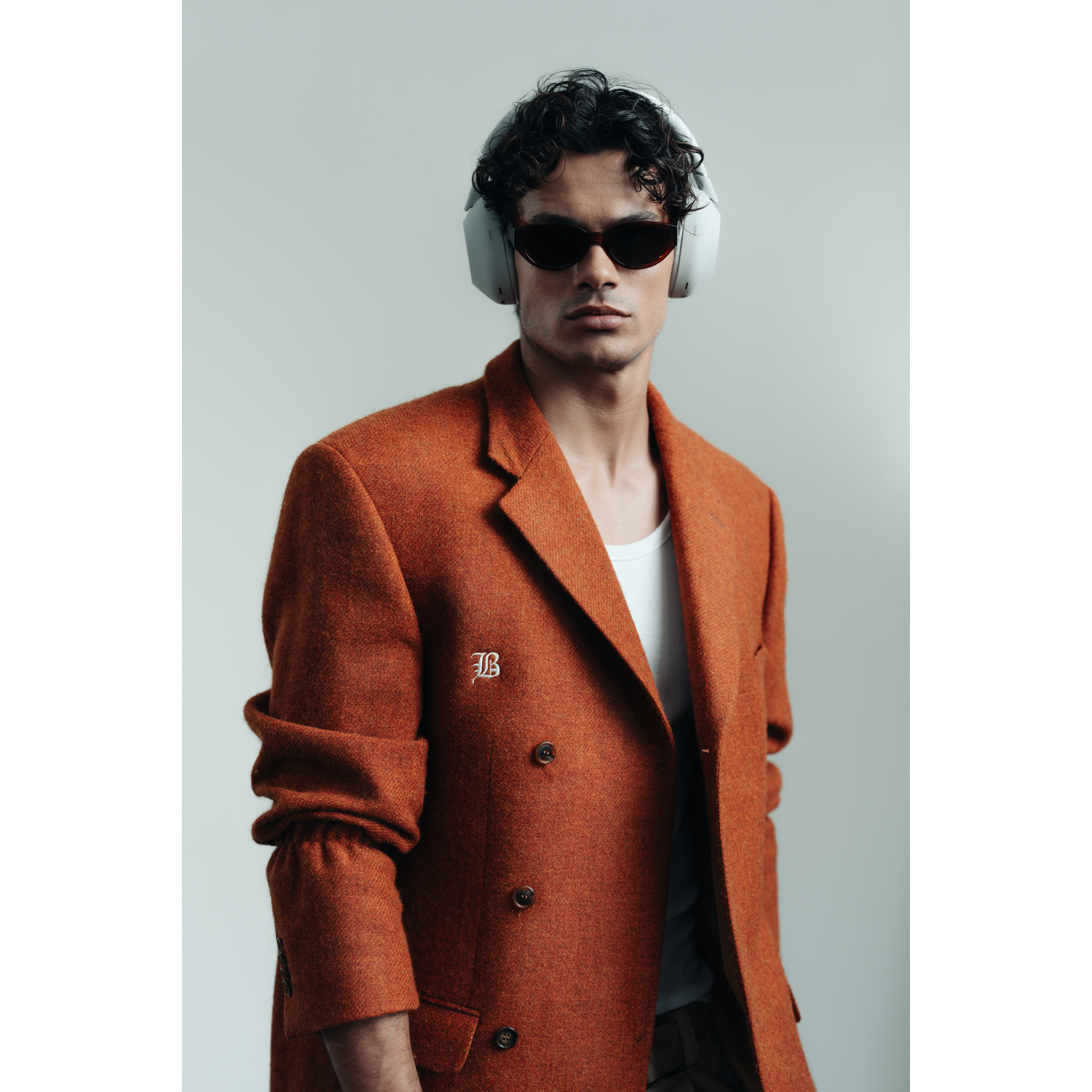 Een man met zonnebril en koptelefoon, draagt een oranje blazer en een wit shirt, tegen een grijze achtergrond.