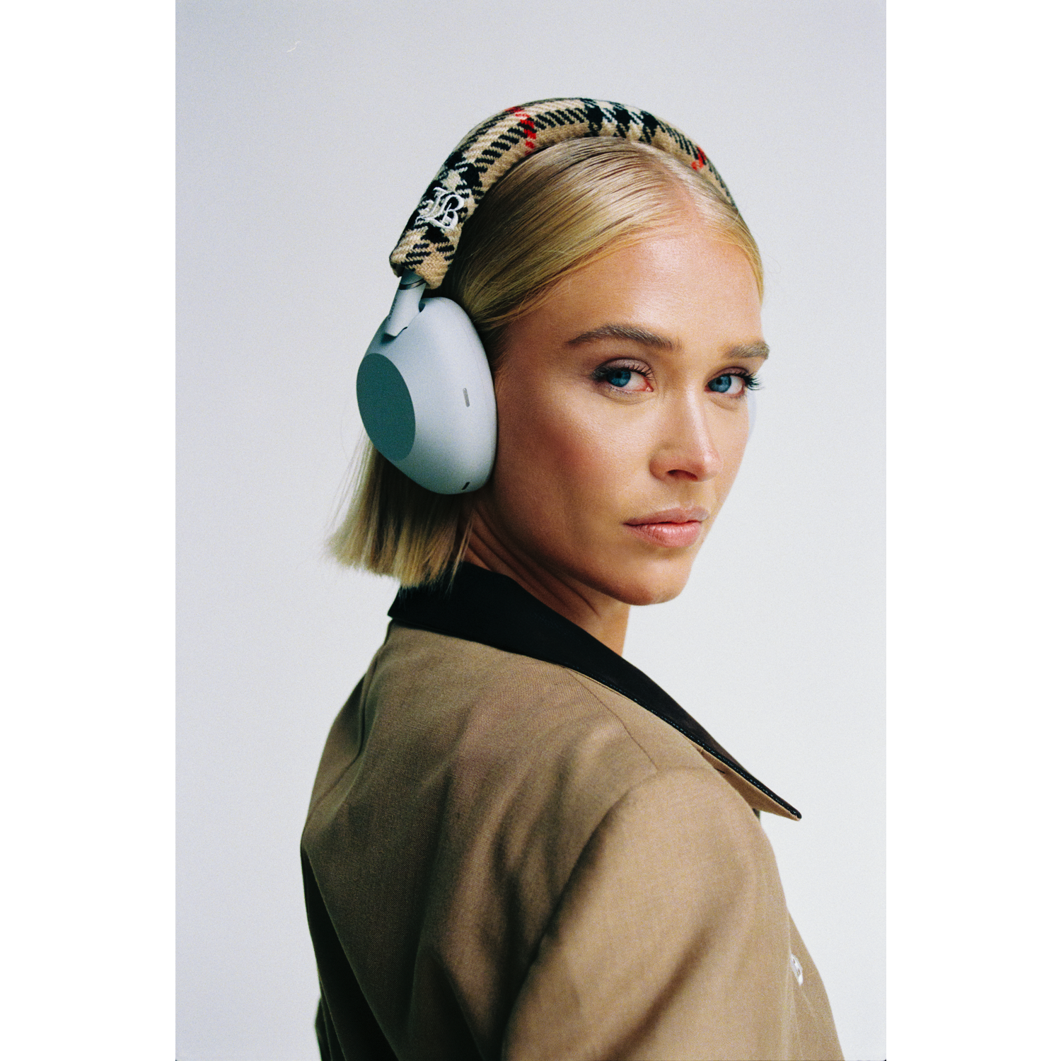 Blonde vrouw met een koptelefoon, kijkt over haar schouder. Ze draagt een beige jasje met een zwarte kraag. Witte achtergrond.