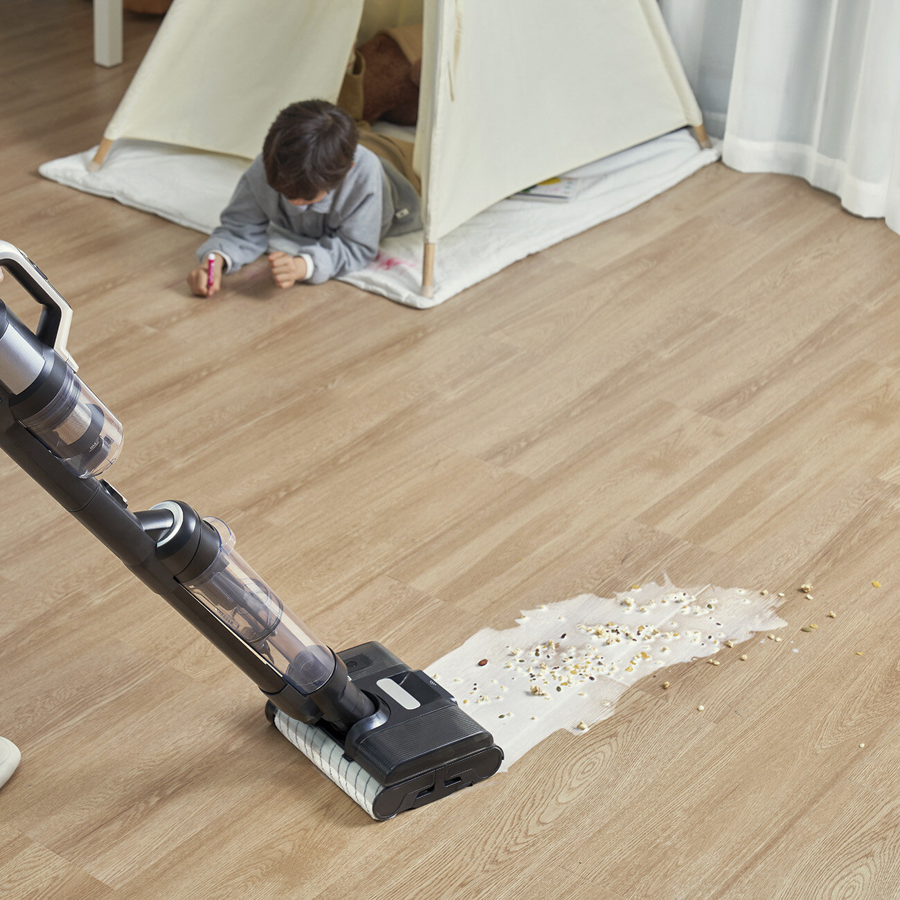 Dziecko jest w namiocie do zabawy, podczas gdy odkurzacz jest używany na drewnianej podłodze do czyszczenia rozlanej cieczy.