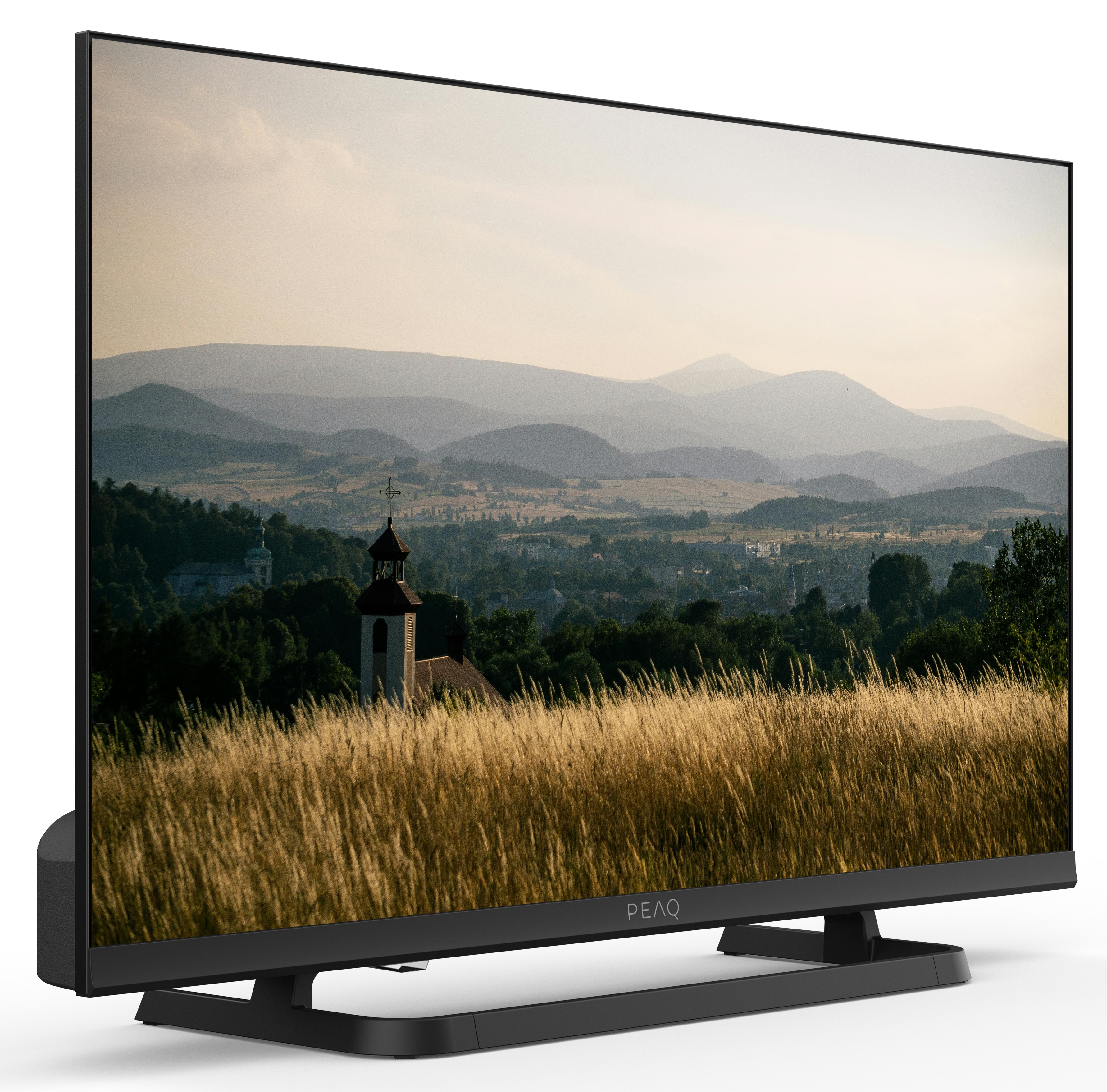 Telewizor wyświetla krajobraz z górami, kościołem i polami. Telewizor jest czarny i stoi na białej powierzchni.