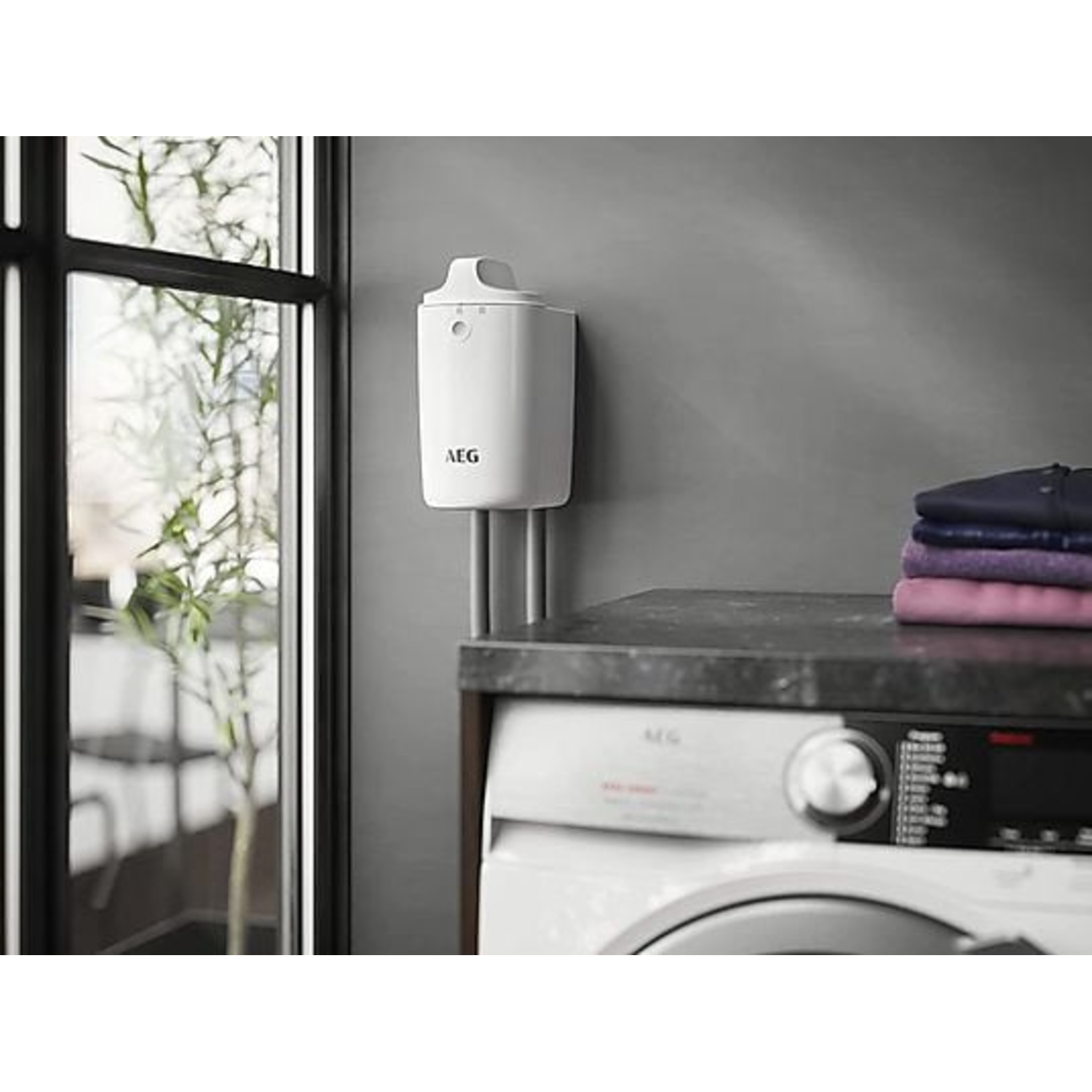 A white AEG appliance is mounted on a gray wall. A washing machine and stacked clothes are below. A window is on the left.