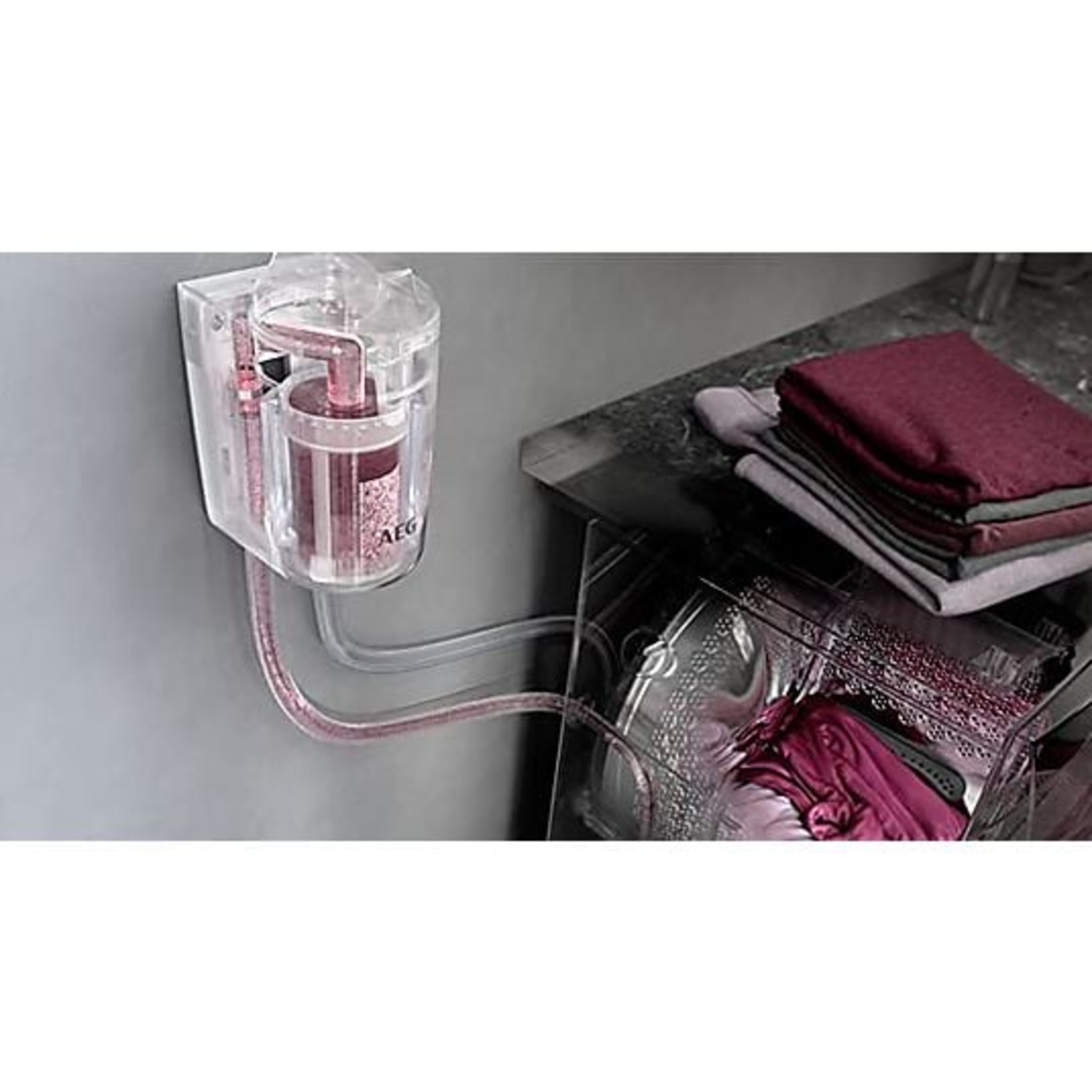 A clear container with pink liquid is attached to a washing machine. Folded clothes are on a counter.