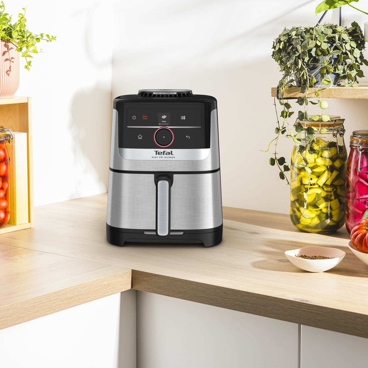 An air fryer sits on a wooden counter. Jars of pickles and plants are in the background.