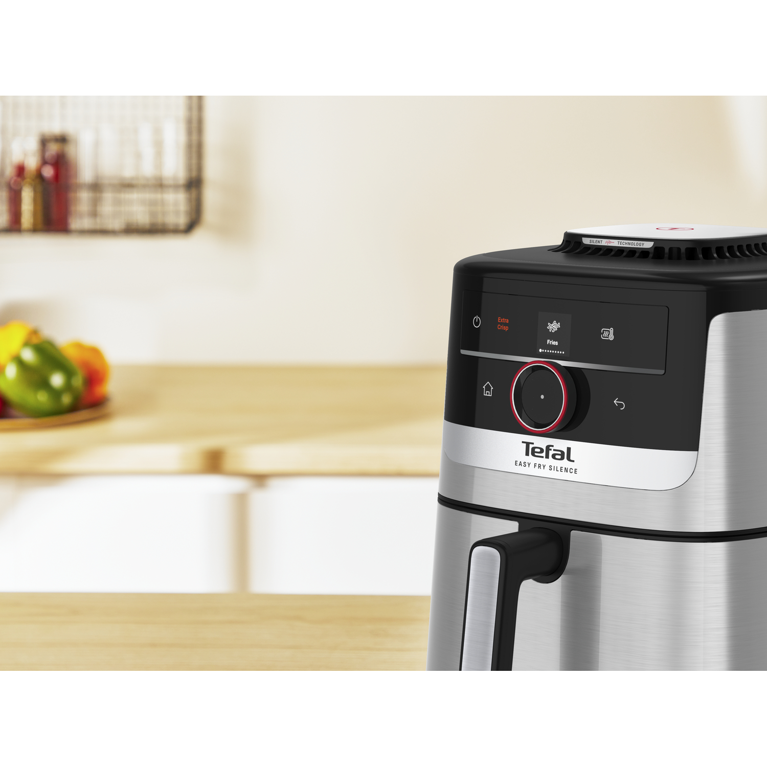 An air fryer with a digital display. It has a black top, silver sides, and a handle. The background shows a kitchen counter and some food.