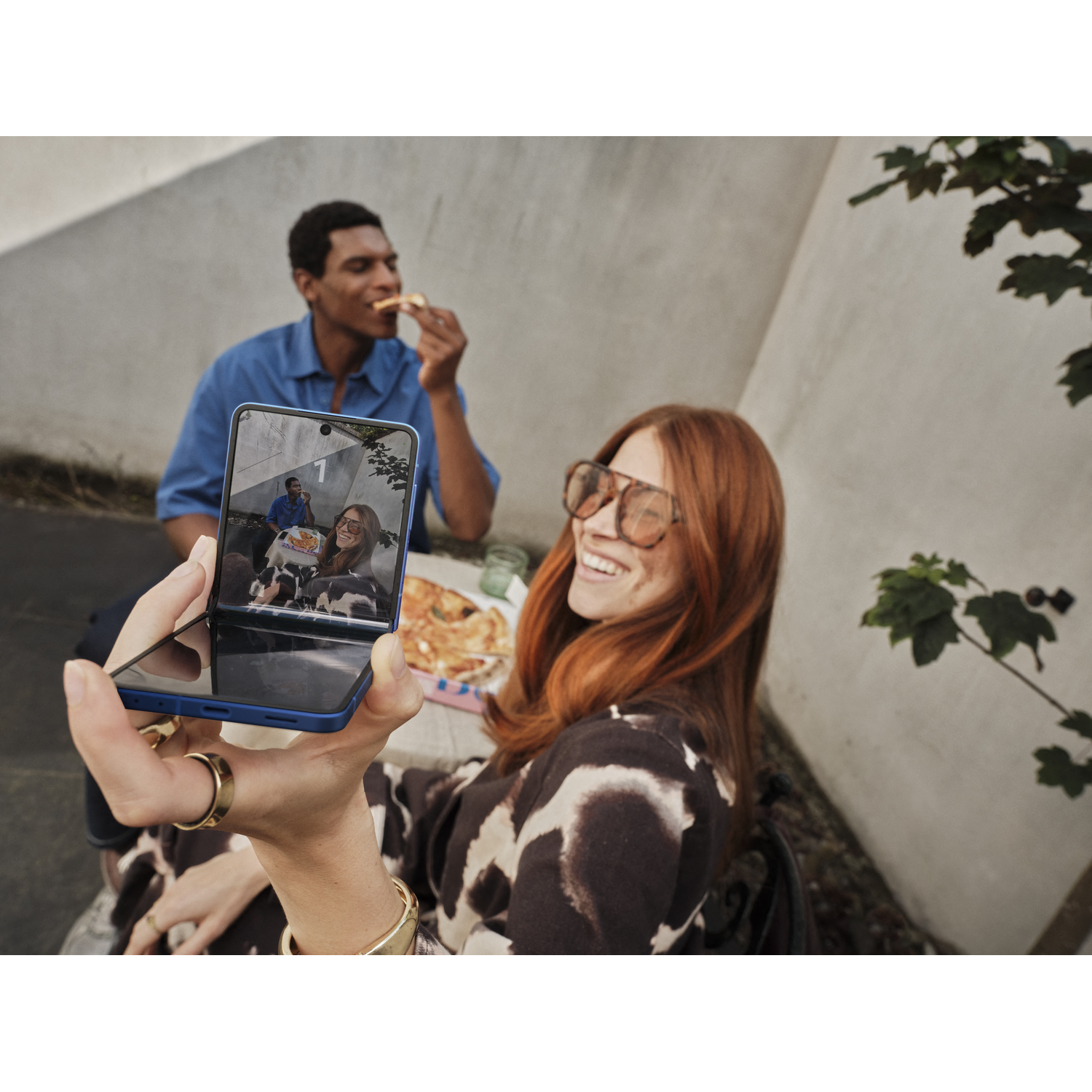 A woman holding a phone, taking a photo of herself and a man eating pizza. She wears glasses and a patterned top.