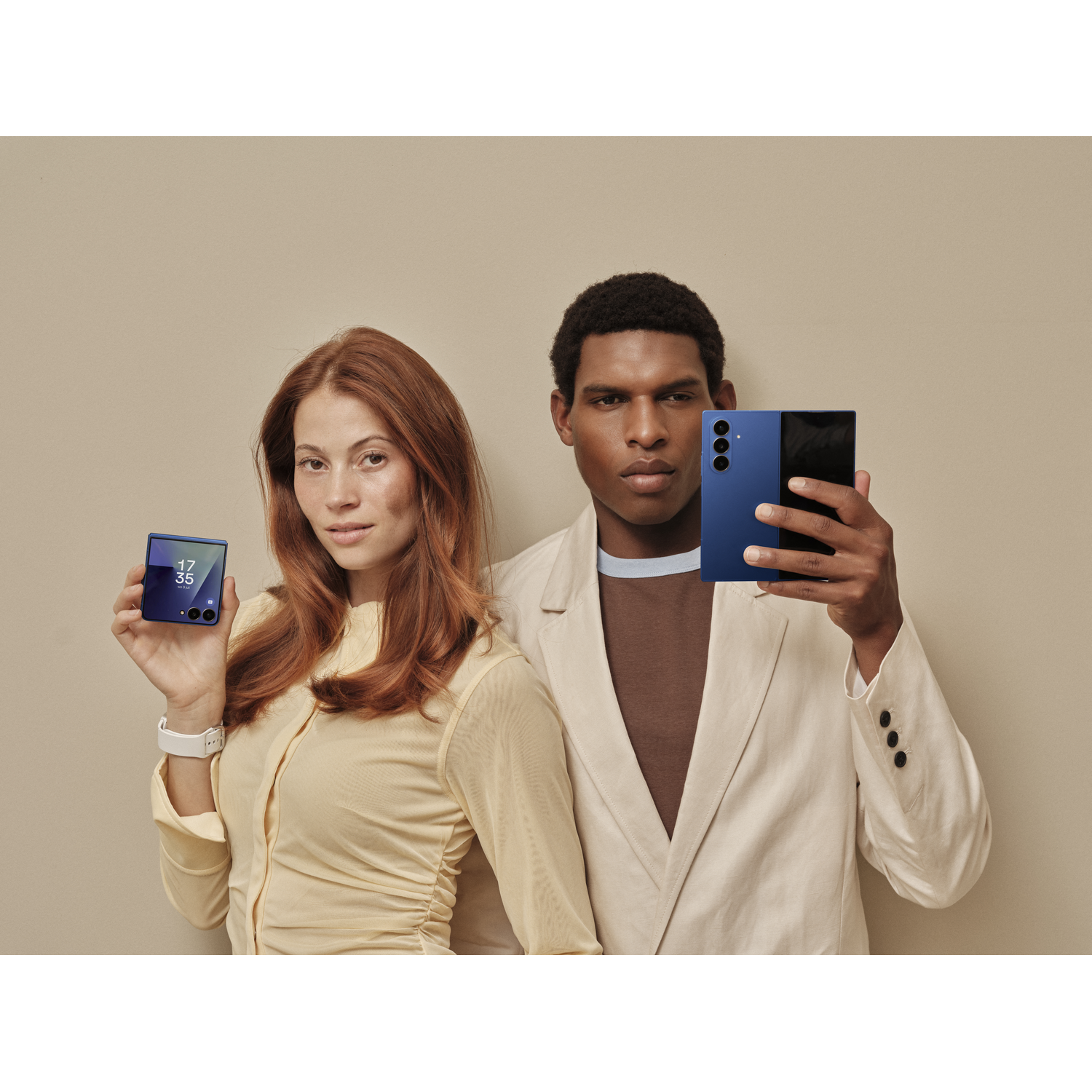 A man and woman hold blue smartphones. The woman has red hair, and the man has dark skin. Beige background.