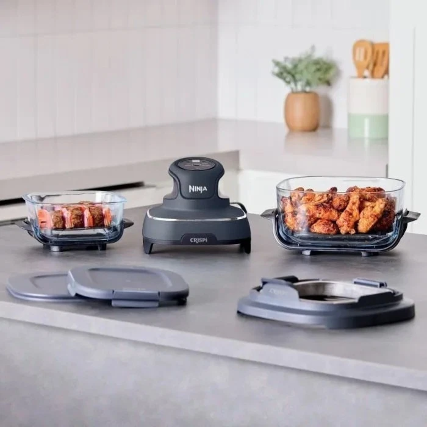 Een Ninja Crispi airfryer met glazen schalen met eten. Deksel en accessoires op het aanrecht.