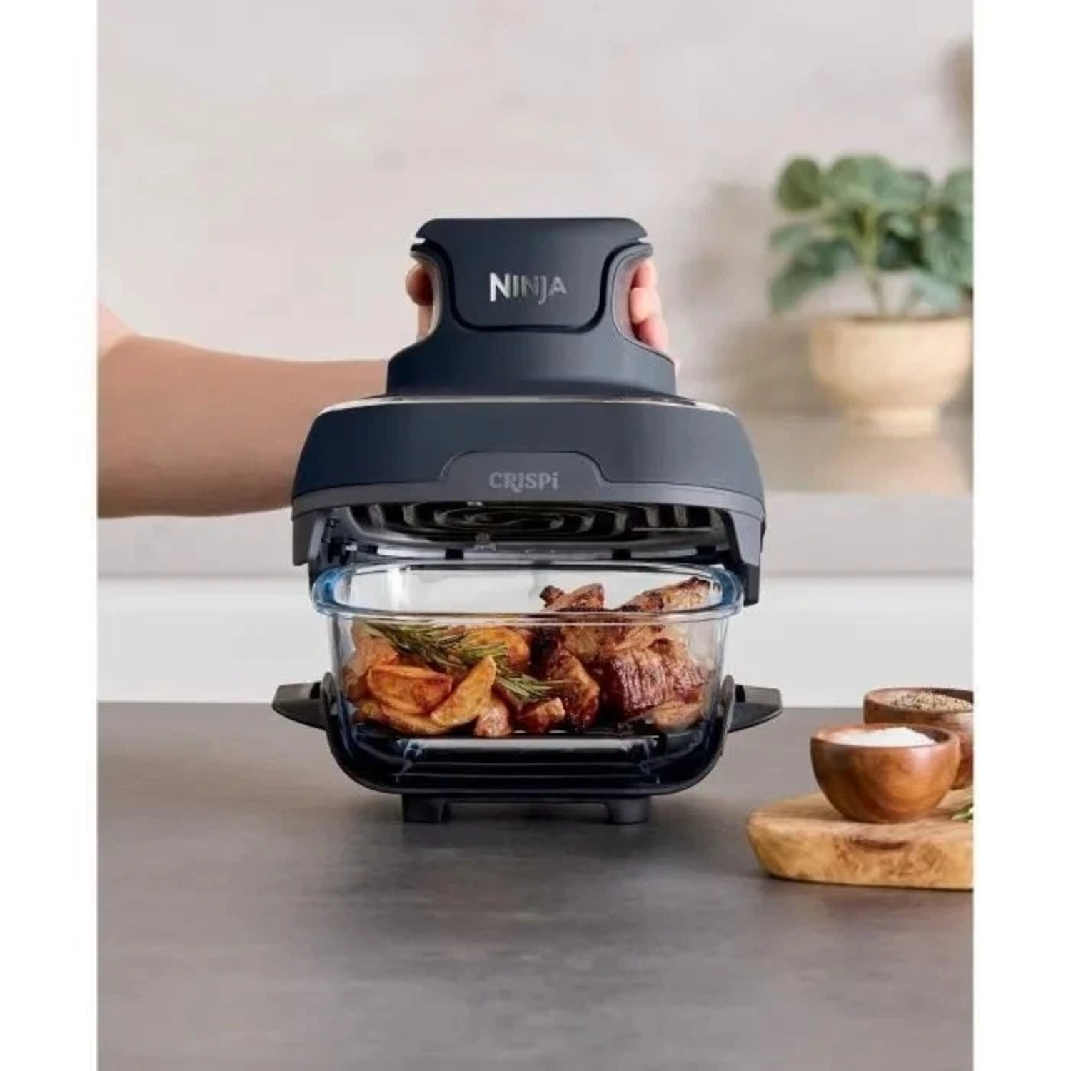 Ninja airfryer met eten erin, vastgehouden door een hand. Aardappelen en vlees zijn zichtbaar. Schalen met kruiden staan op een houten plank.