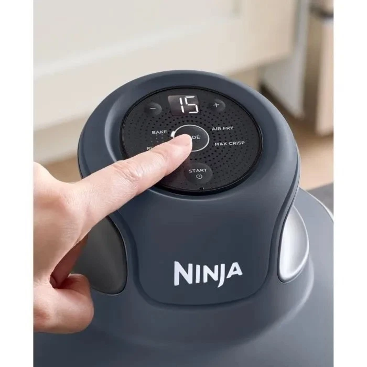 Een hand drukt op de 'mode'-knop van een Ninja-apparaat. Het display toont 15. Andere knoppen zijn 'bakken' en 'airfryer'.