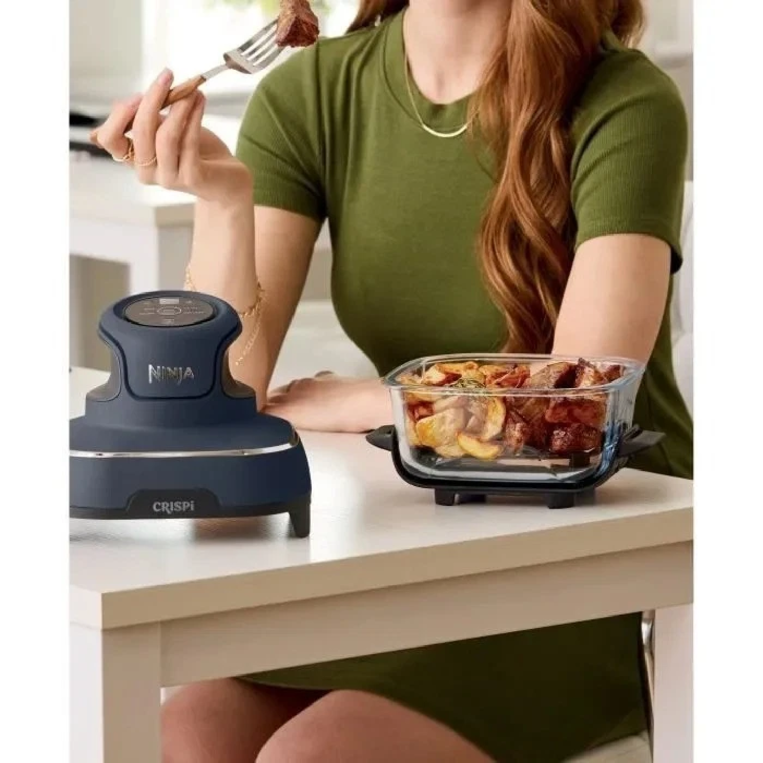Een vrouw in het groen houdt een vork met eten vast. Een Ninja Crispi-apparaat en een glazen schaal met eten op een witte tafel.