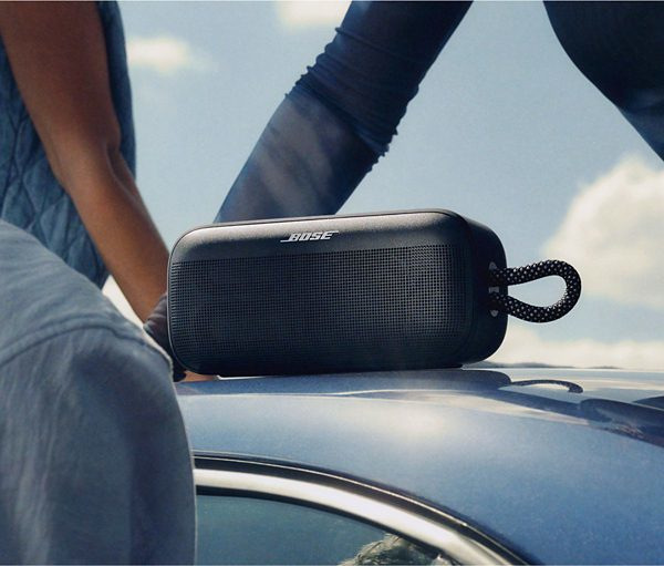 Czarny głośnik Bose spoczywa na samochodzie. Ludzie w dżinsach są w tle. Niebo jest niebieskie.